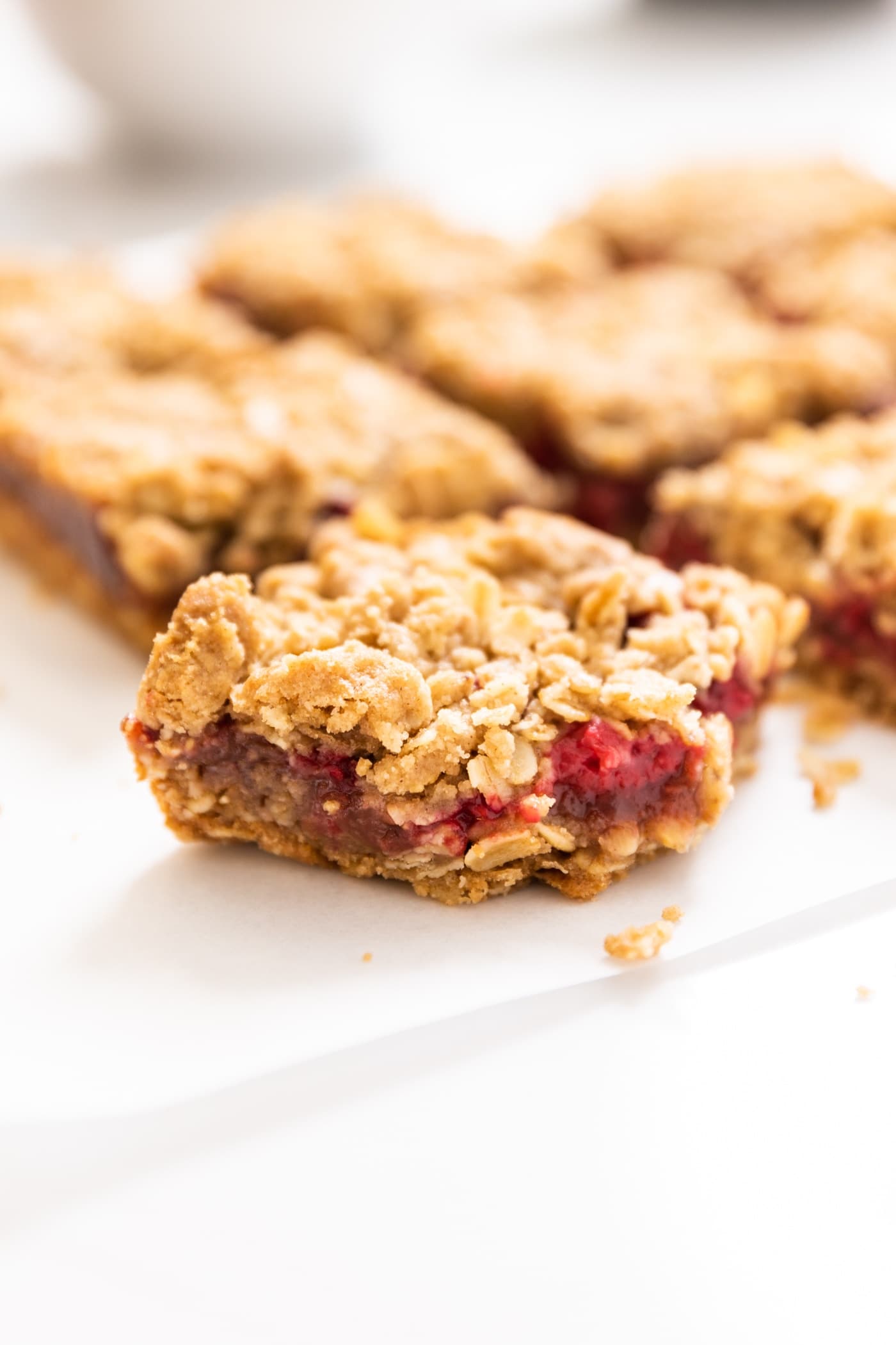 One square raspberry crumb bar sitting on white piece of parchment with a bit of red colored filling oozing out with extra bars in background
