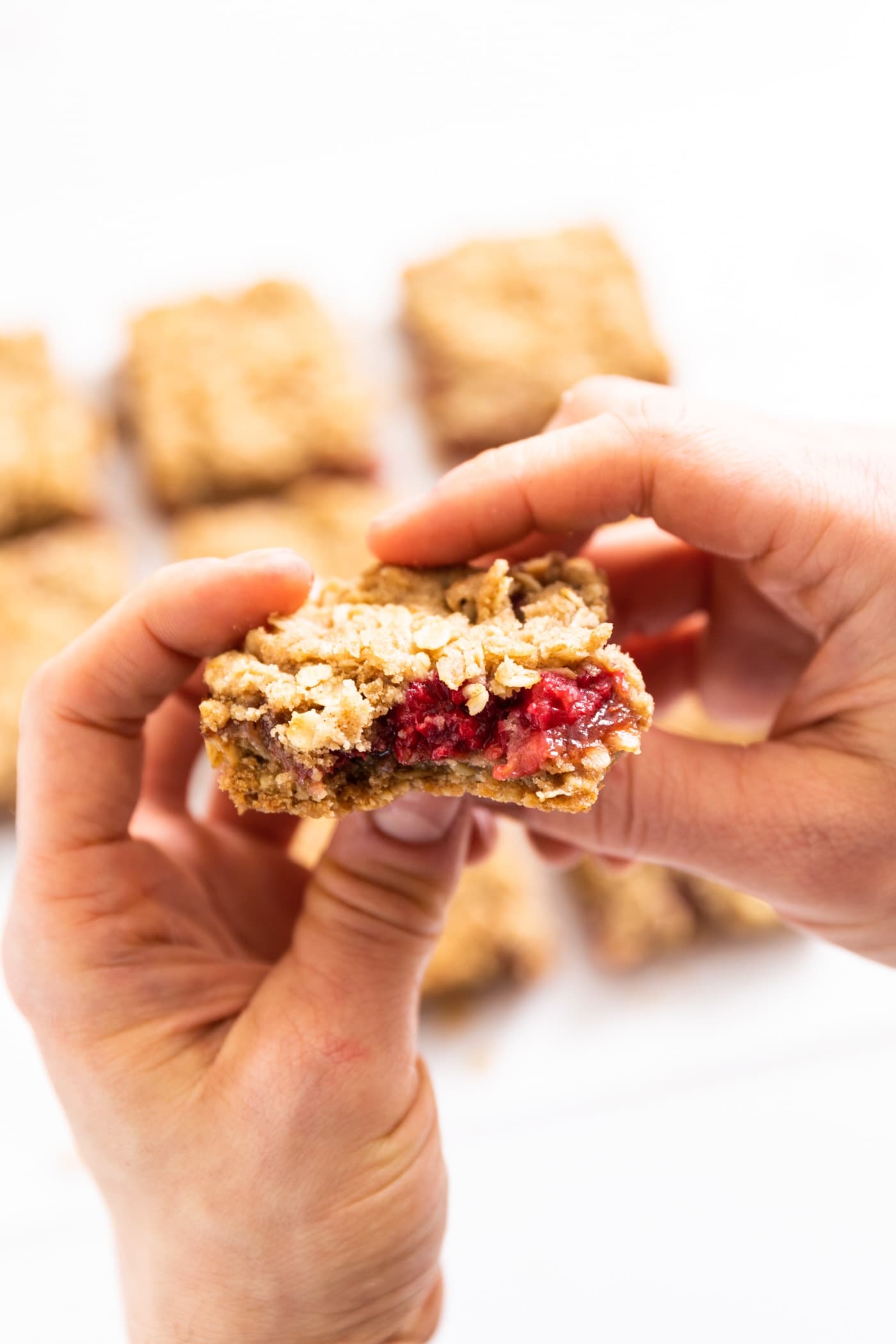 Two hands holding a raspberry crumb bar with a bite taken out with extra square bars in the background all on a white surface