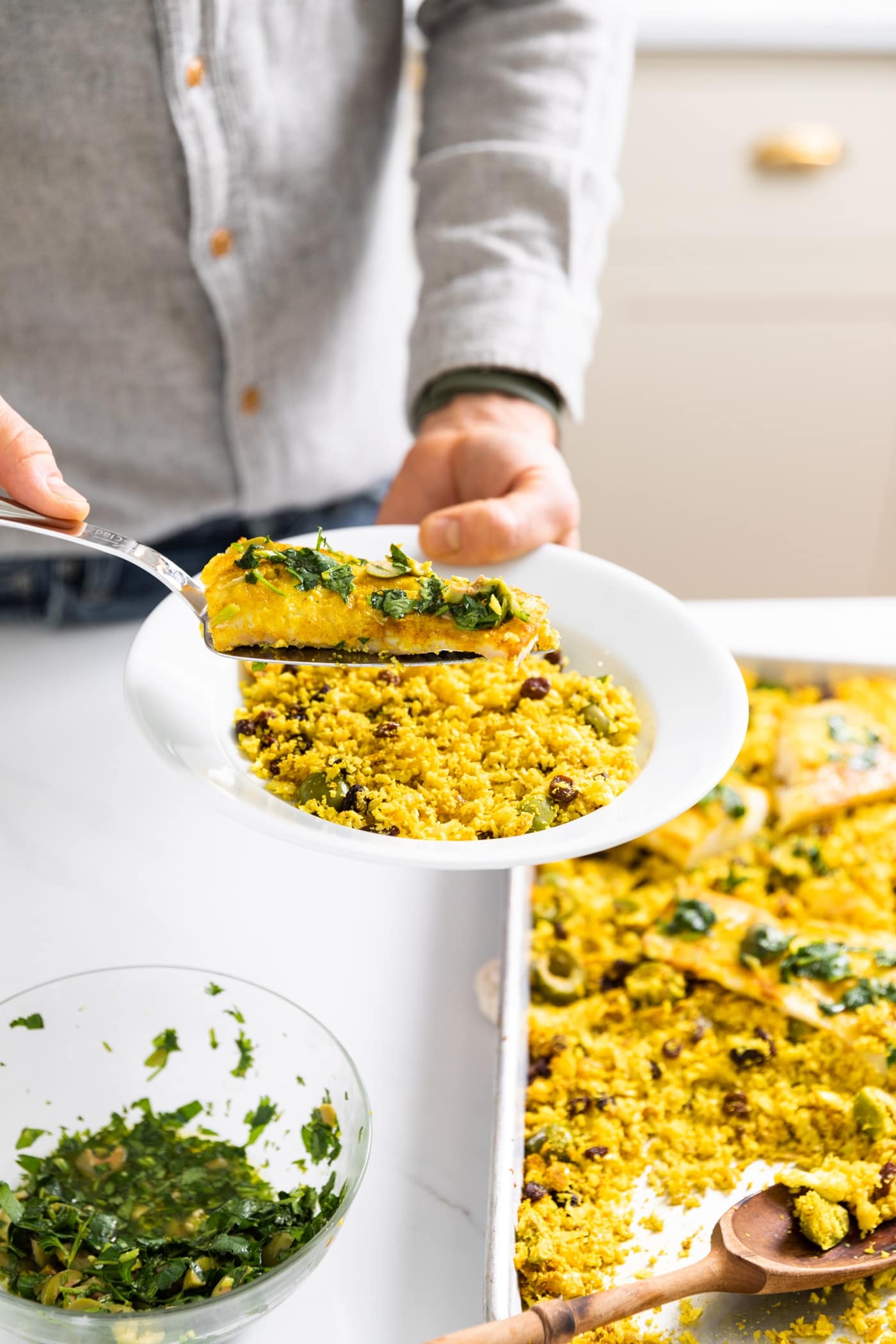 White plate held by man wearing a gray button down shirt putting piece of fish onto bed of yellow colored cauliflower rice