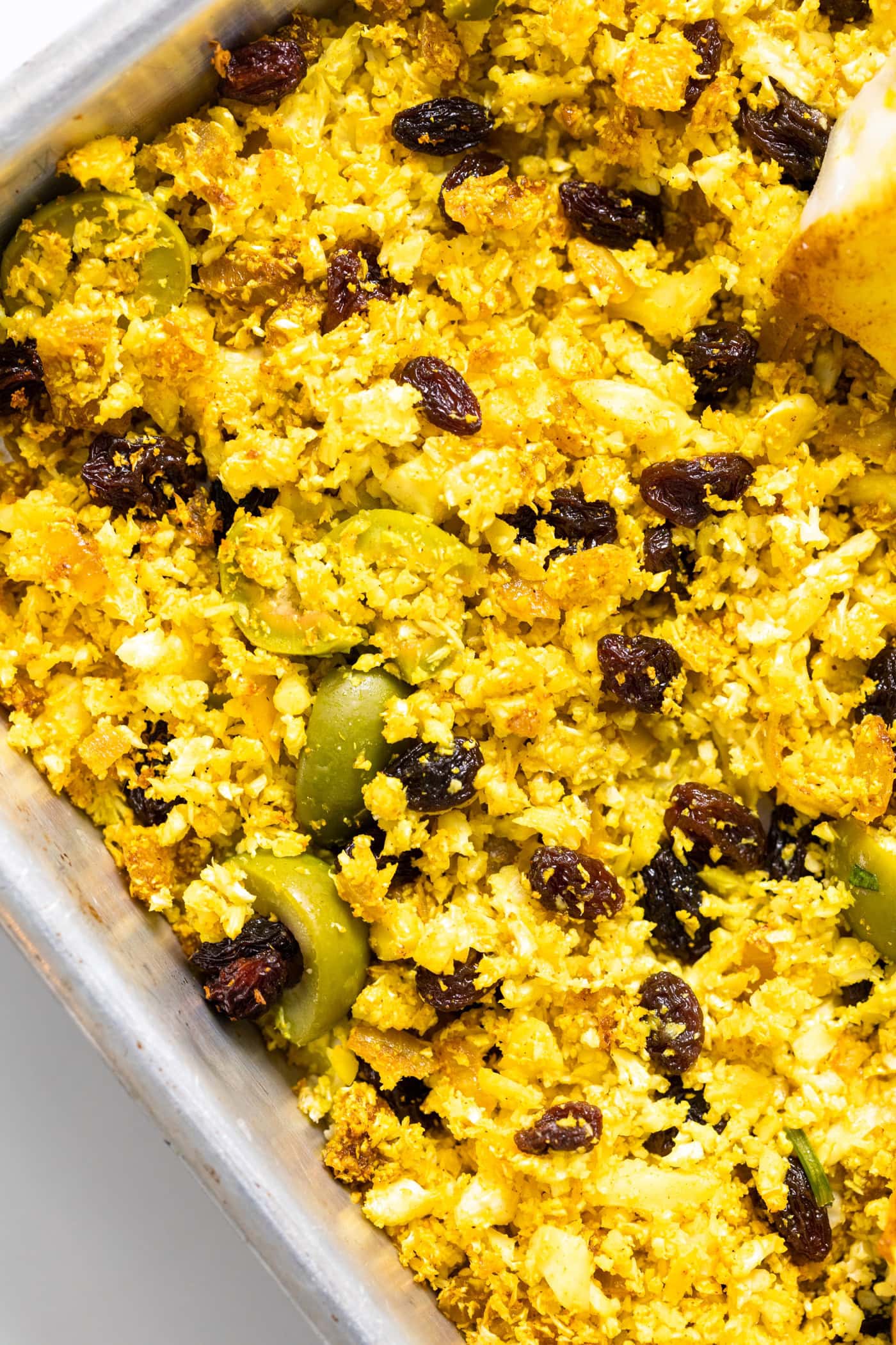 Top down view of yellow colored cauliflower rice with pieces of olives and raisins mixed amongst all on sheet pan