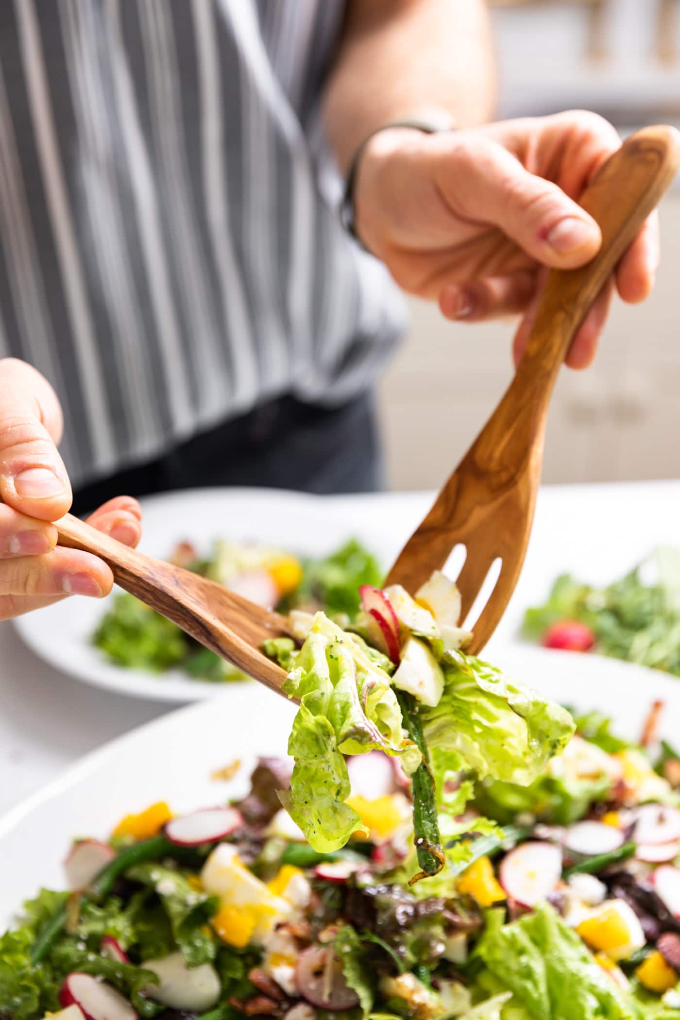Two hands holding wooden salad tongs lifting up pieces of green lettuce with platter filled with rest of salad below