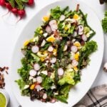 Top down view of white serving platter filled with green butter lettuce topped with fresh spring vegetables and hardboiled egg with extra ingredients sitting all around on white countertop
