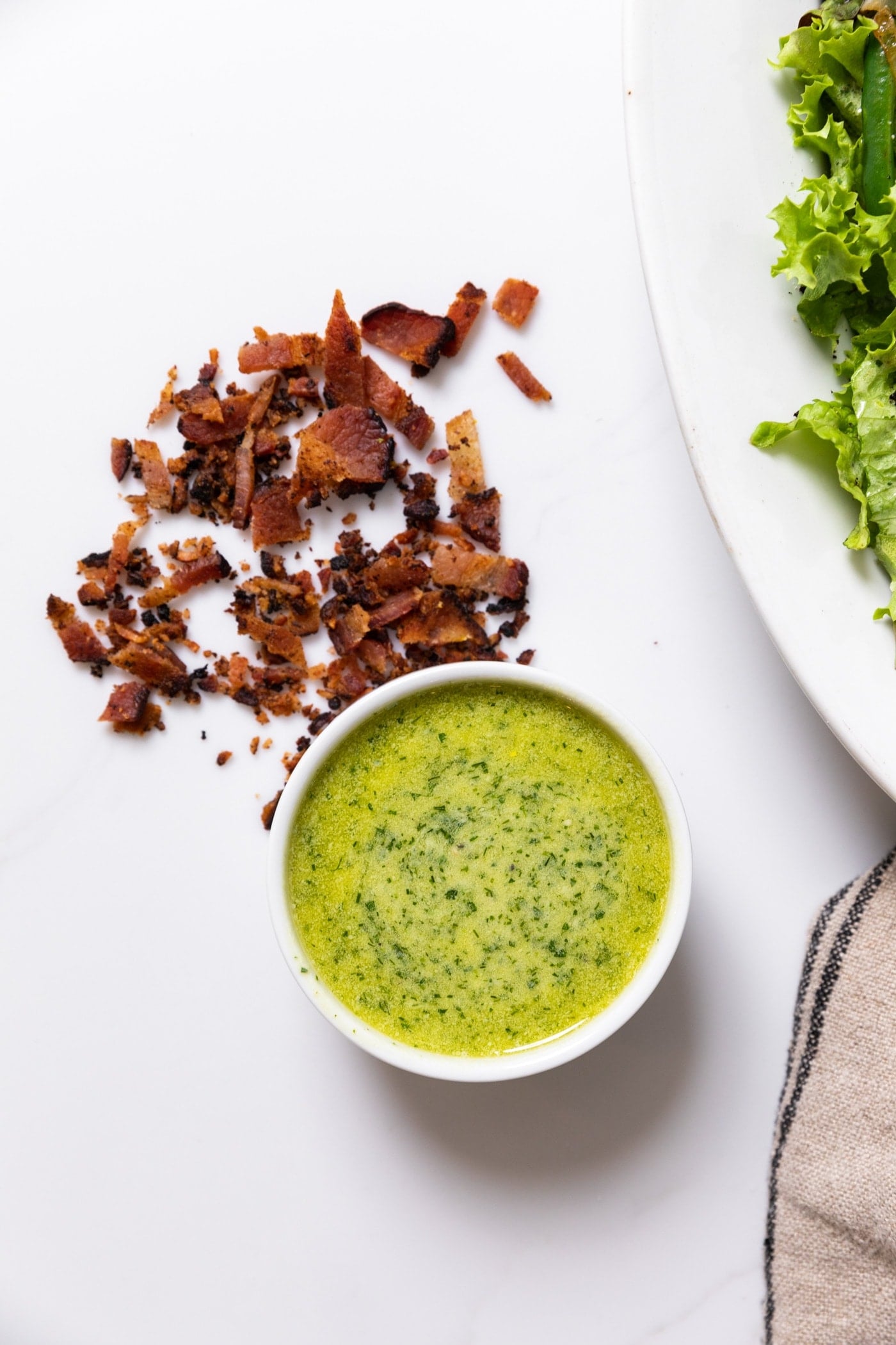 Top down view of round white bowl filled with green salad dressing liquid with chopped bacon to the side all on white countertop