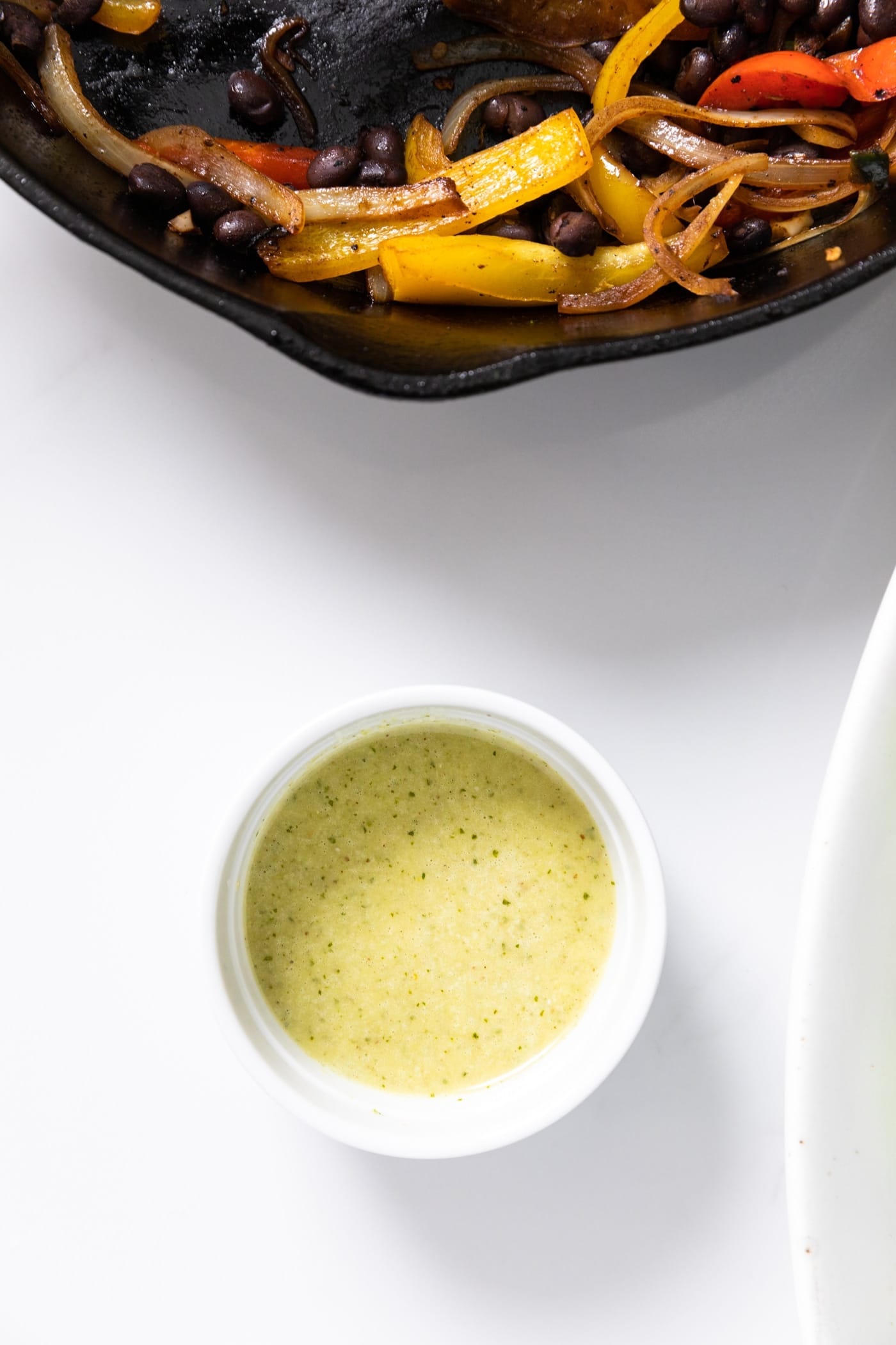 Top down view of white countertop with small white bowl filled with light green salad dressing sitting on top with fajita vegetables above