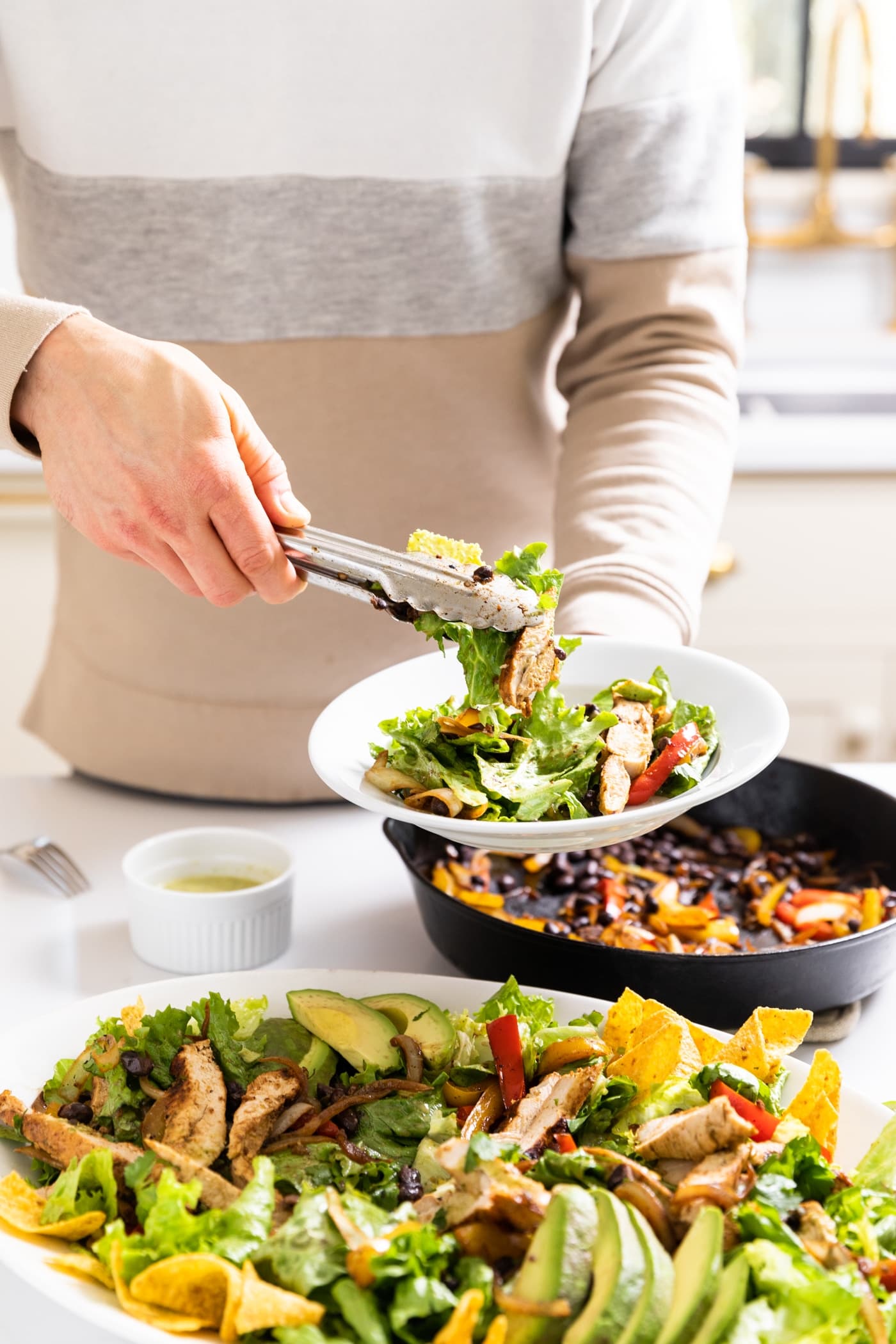 Hand holding tongs putting salad on white serving plate with rest of salad in platter below with black skillet filled with peppers in background