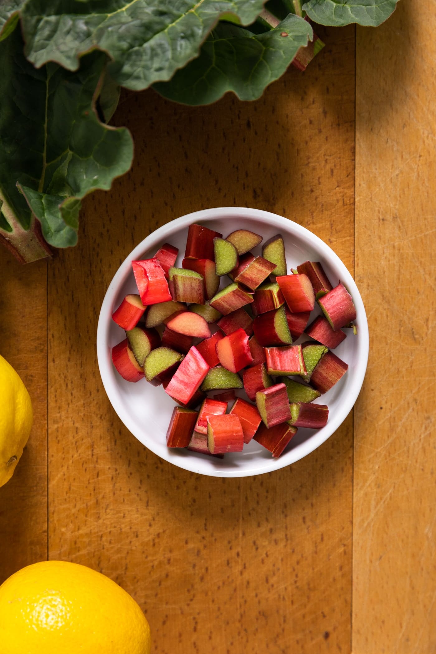 Top down view of chopped rhubarb sitting on wooden board with rhubarb leaf off to the side along with lemon