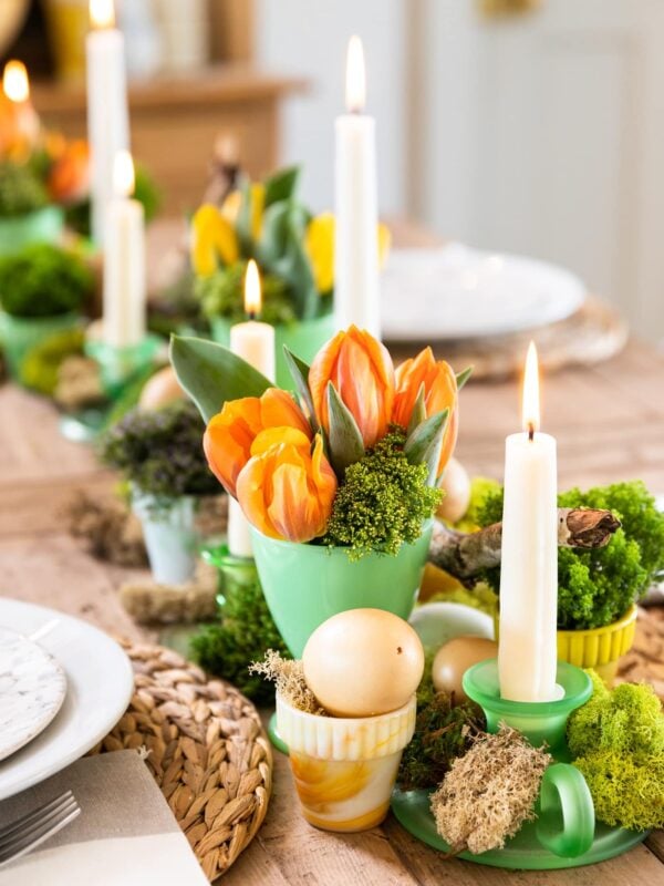 Green vase filled with orange tulips all on wood table with candles lit and white plates set around