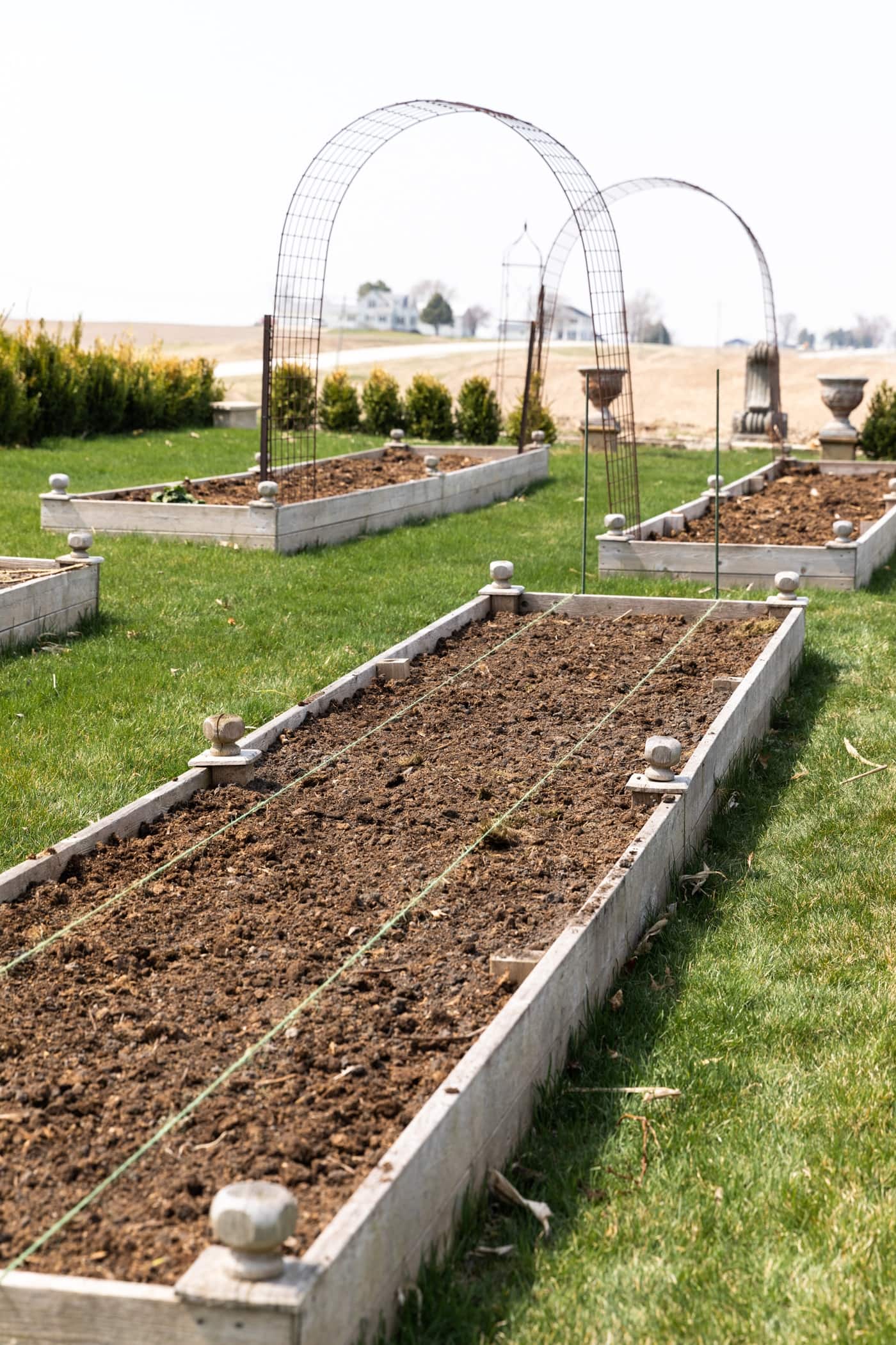 Raised garden bed filled with dirt surrounded by green grass with arches in distance