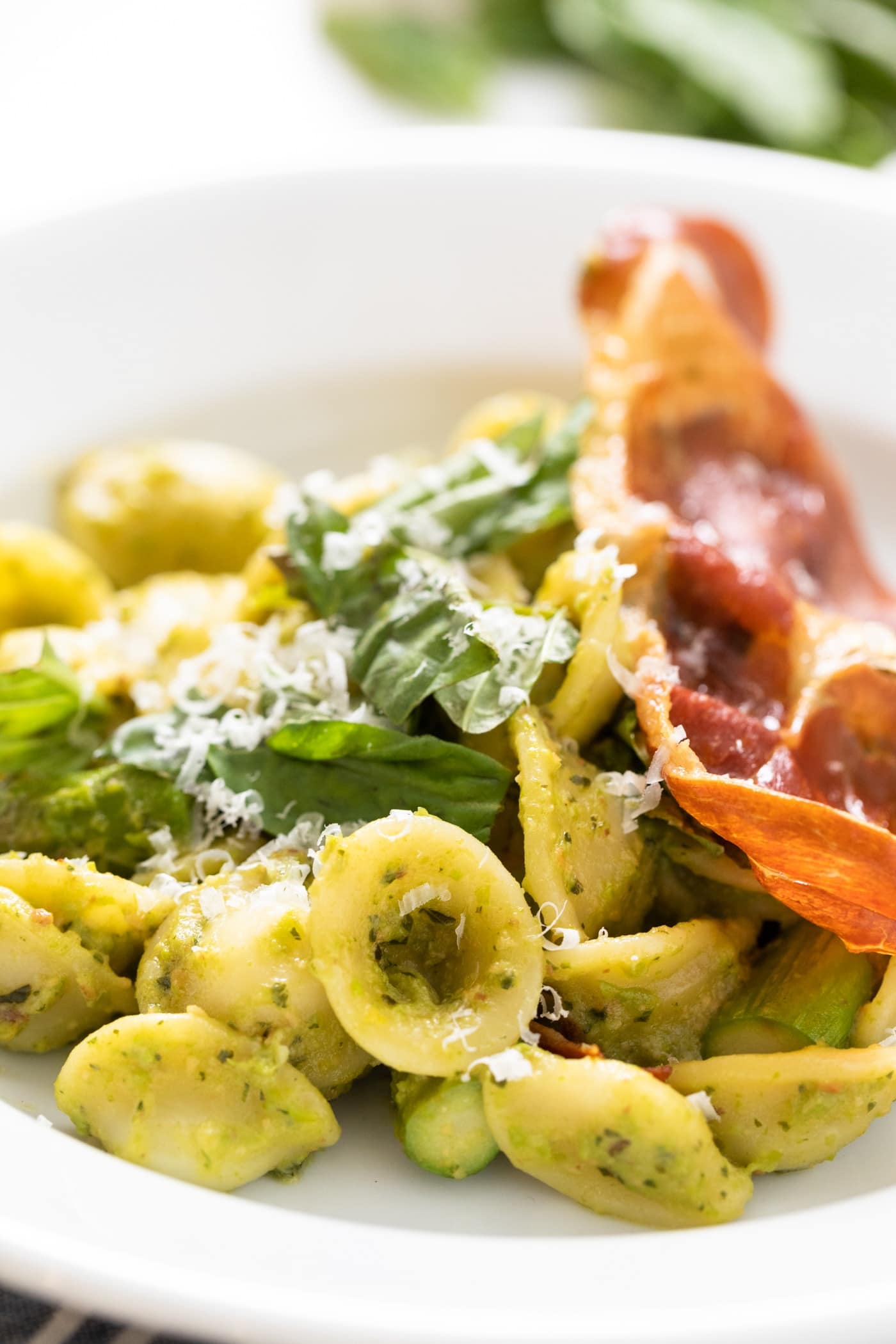 Close up view of pieces of pesto covered pasta sitting on white plate with slice of crispy prosciutto and basil leaves sitting on top