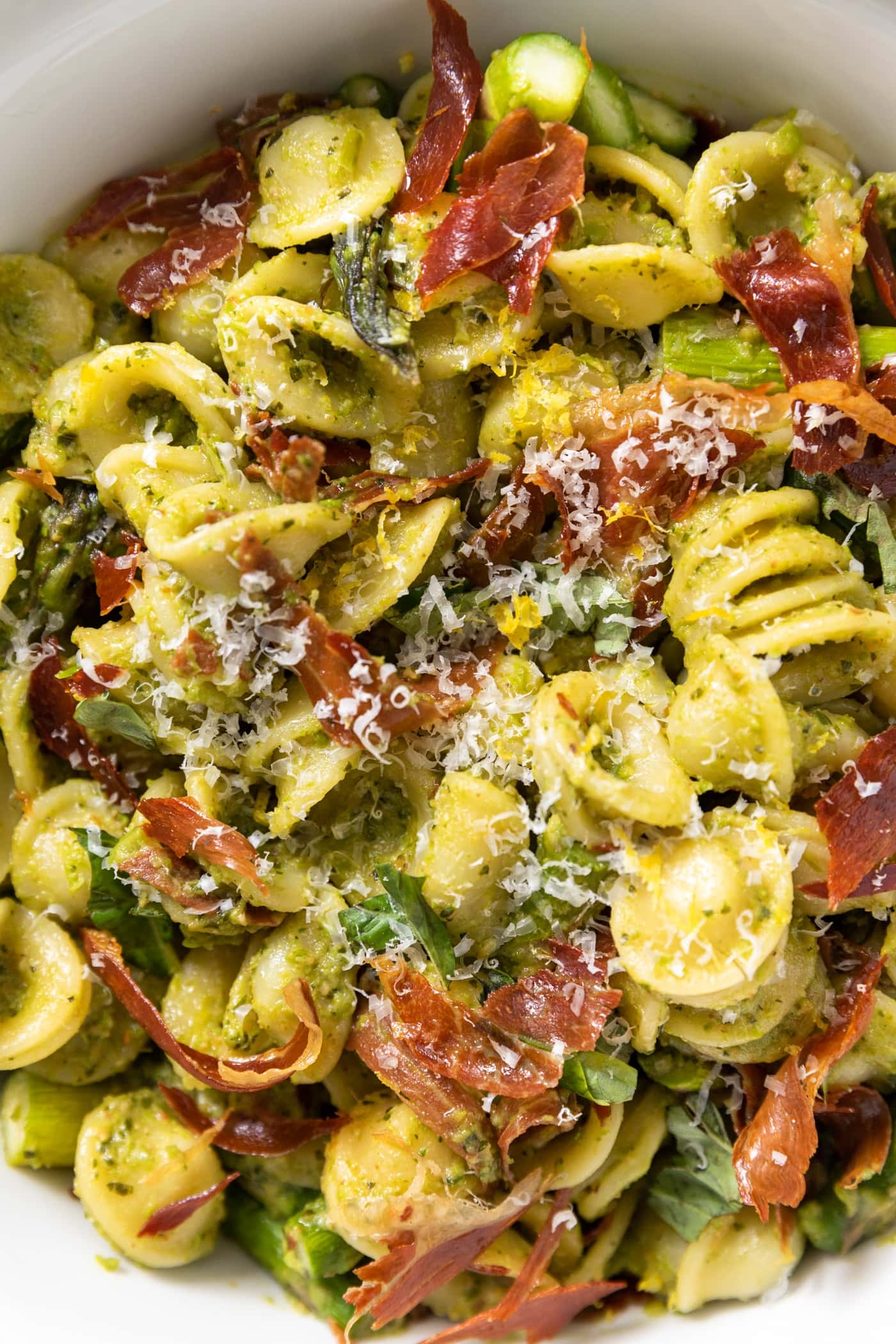 Close up view of pieces of yellow colored pasta mixed with asparagus and pieces of crispy prosciutto all in large white bowl