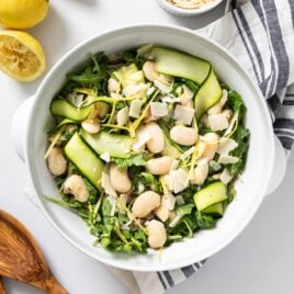 Top down view of large white bowl filled with greens and white beans topped with lemon zest sitting on white countertop