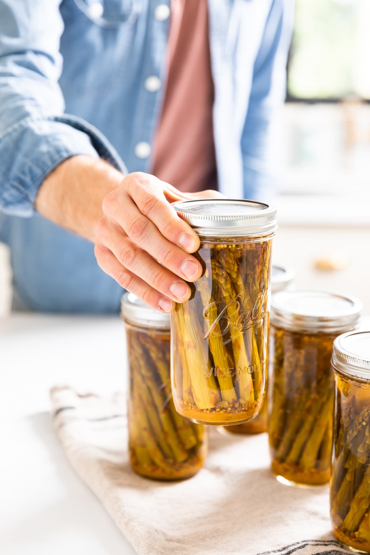 Hand holding glass jar with spears of pickled asparagus with other jars sitting around all on white countertop