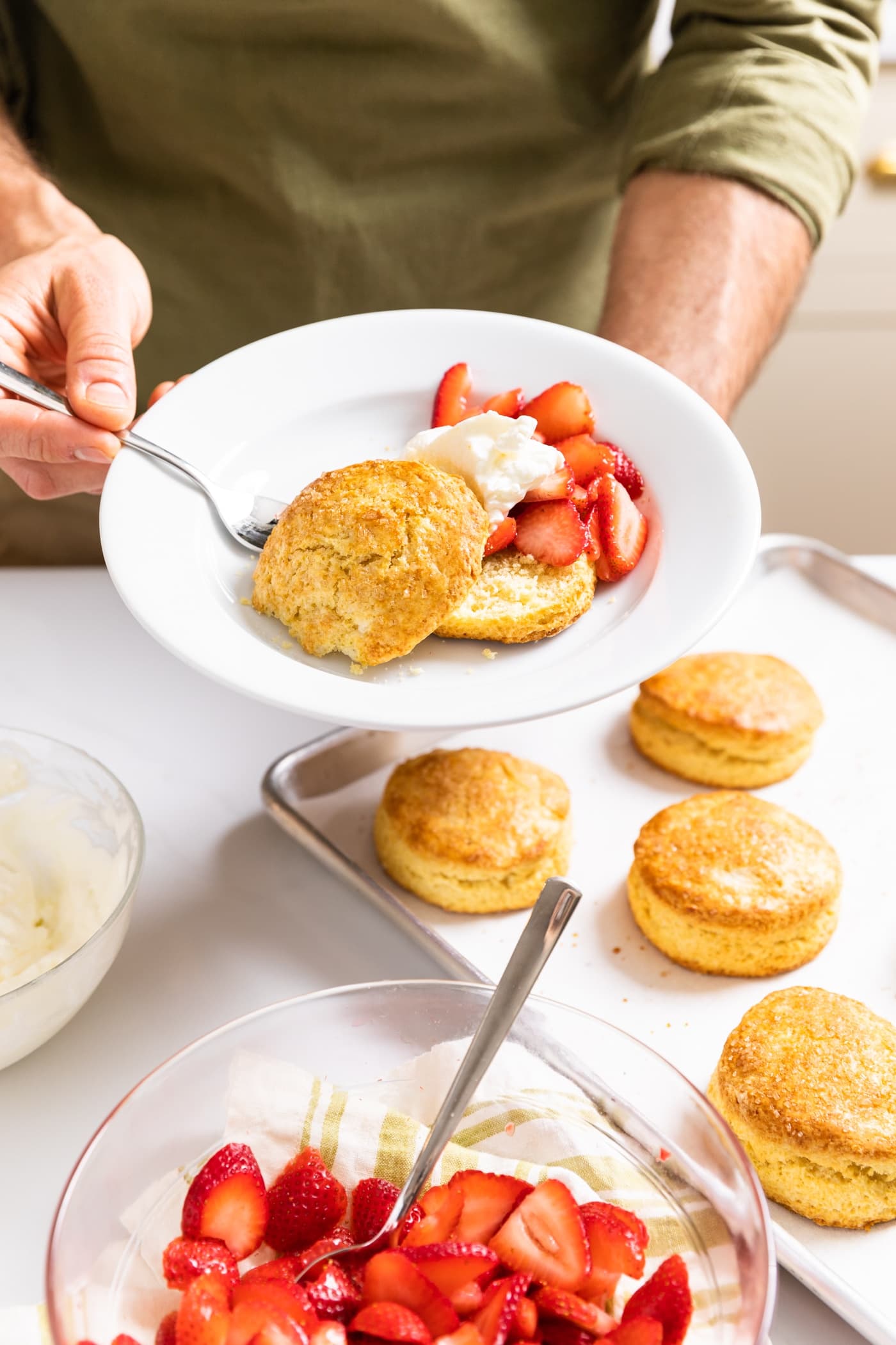 Two hands holding a white plate with yellow biscuit and strawberries topped with whipped cream with rest of components sitting around on countertop