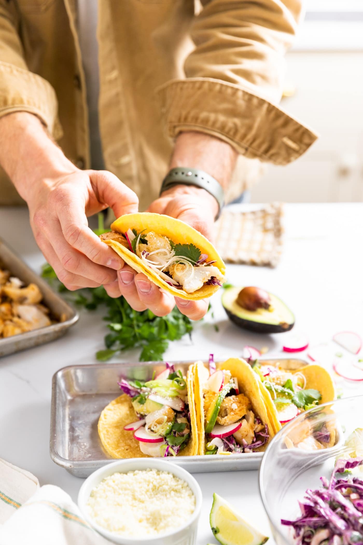 Two hands holding a yellow colored tortilla filled with cod with extra tacos sitting on countertop with toppings all around