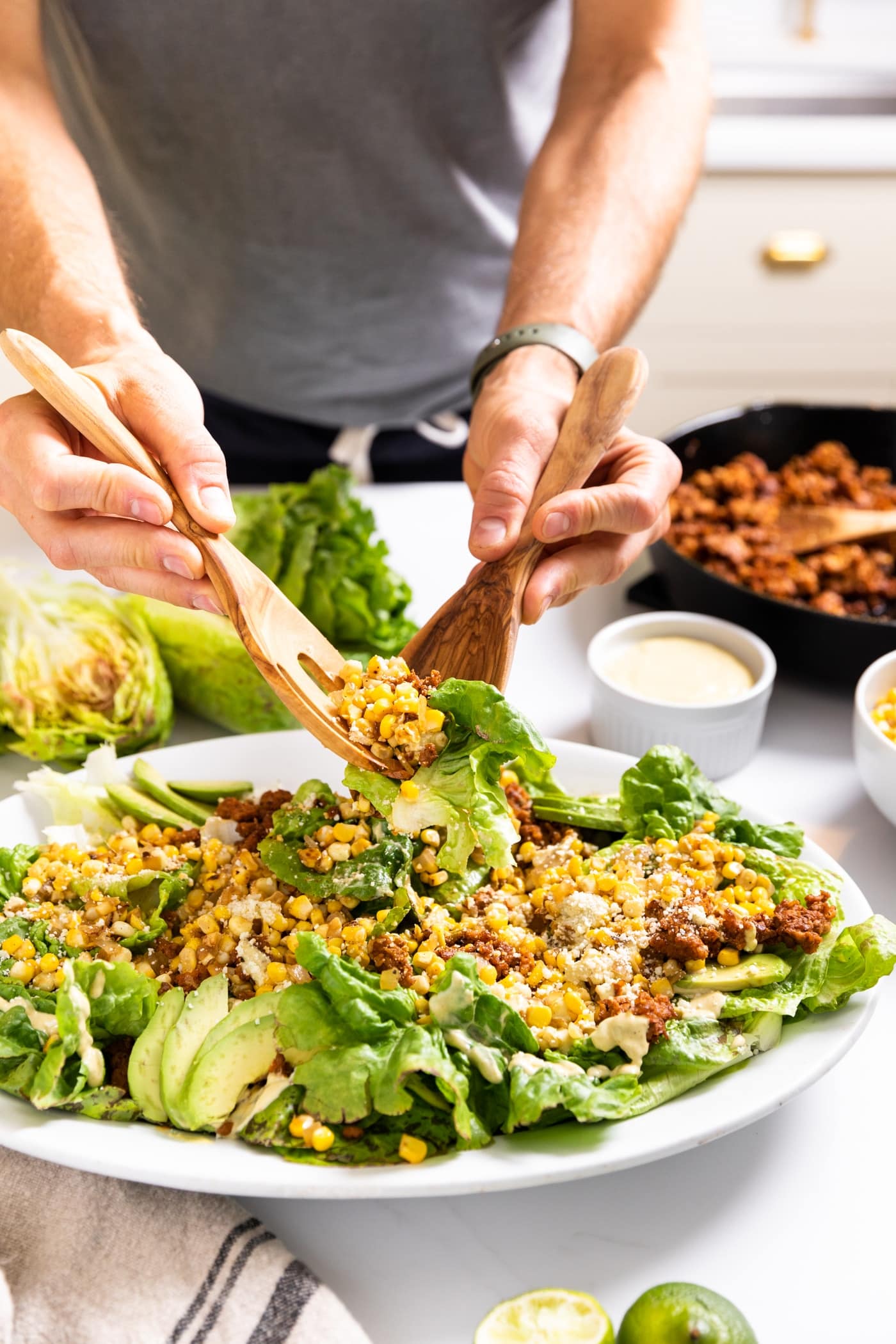 Two hands holding serving tongs full of salad packed with sweet corn and chorizo and lettuce with extra toppings in background
