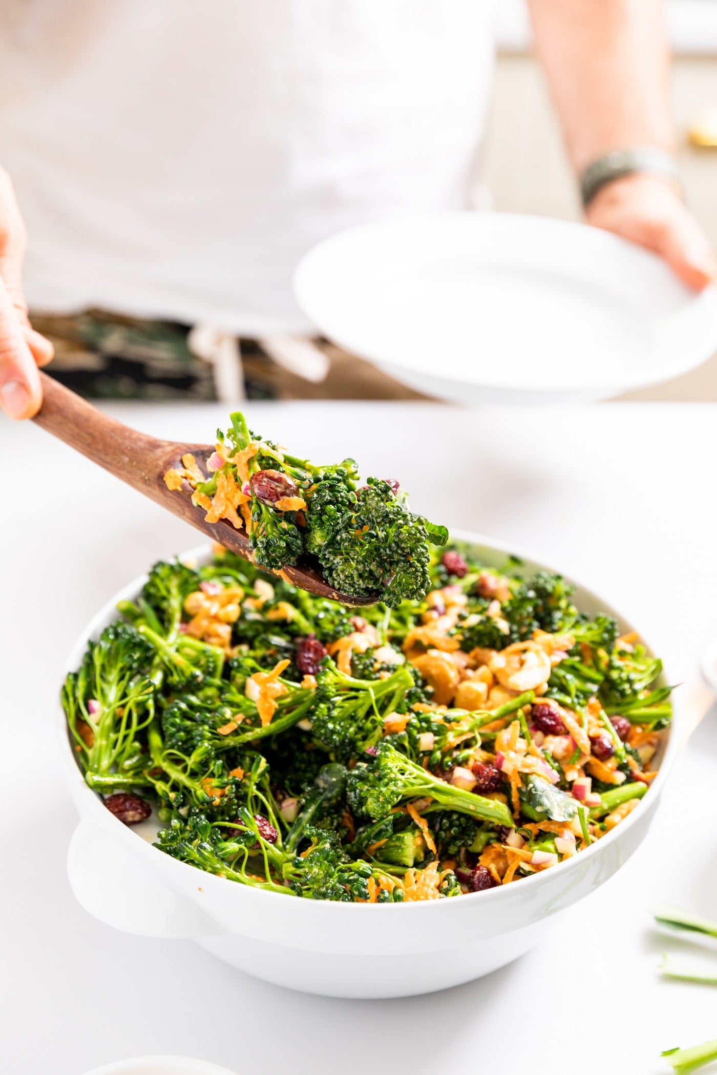 White bowl filled with broccoli florets topped with sauce and cashews and dried cranberries with hand holding wooden serving spoon