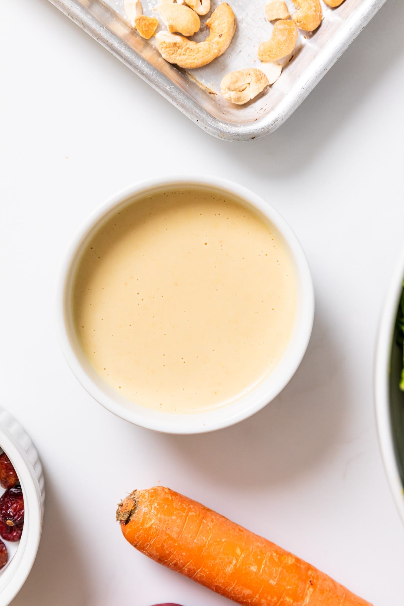 Top down view of small white bowl filled with taupe colored dressing with cashews and carrot sitting around all on white countertop