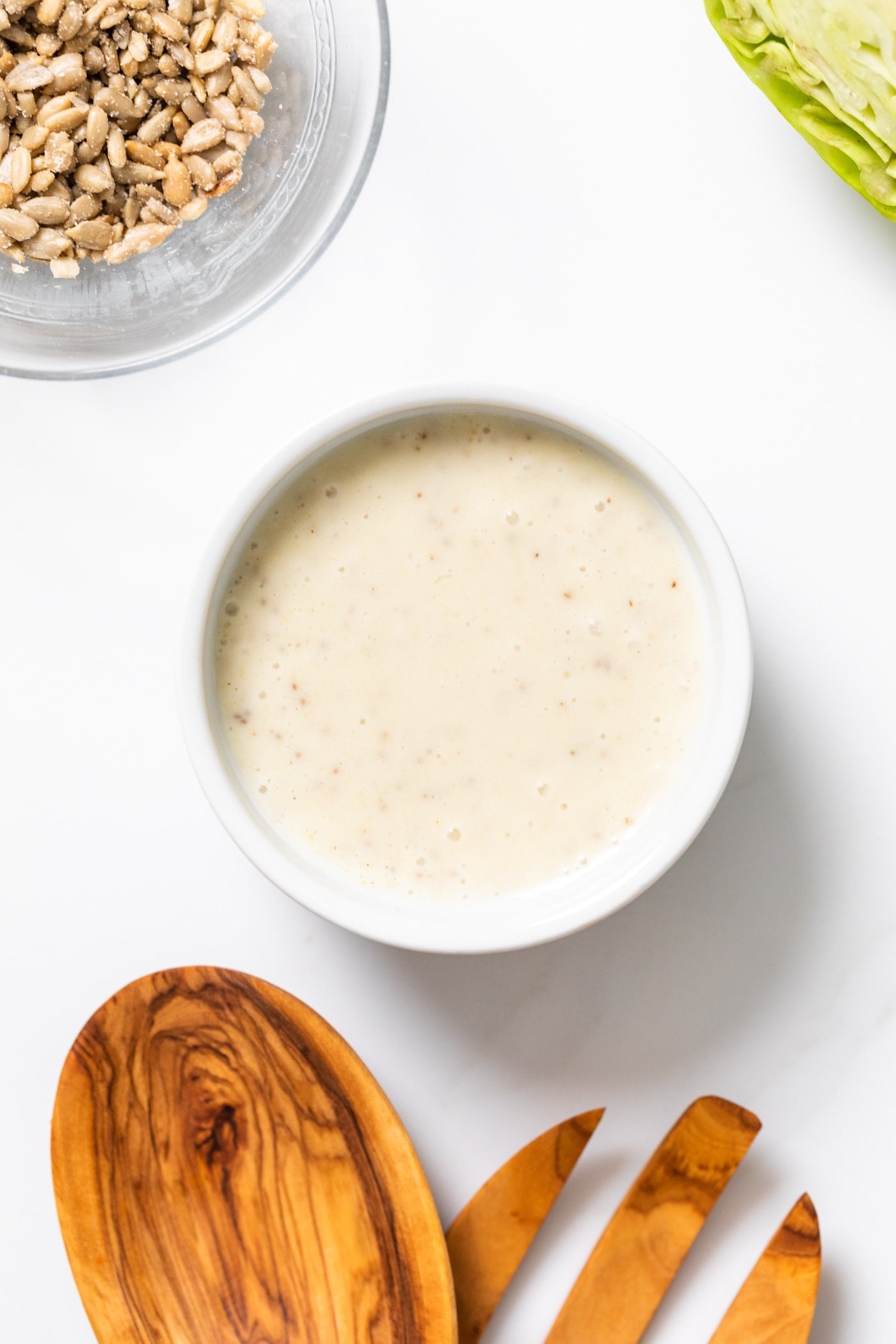 Top down view of white colored salad dressing sitting on white countertop with wooden tongs