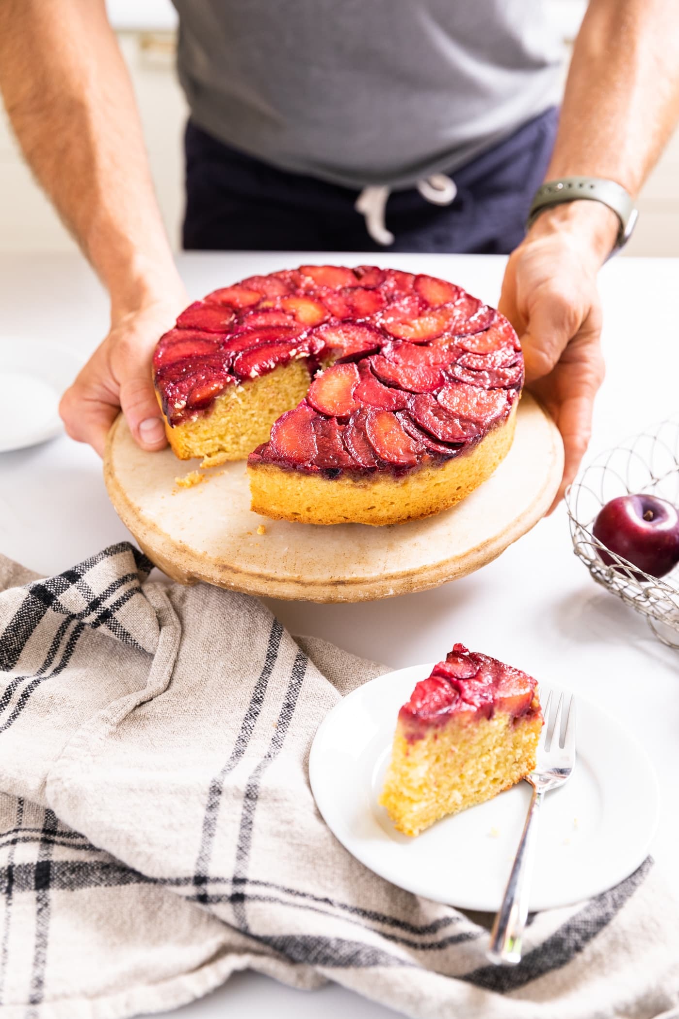 Two hands holding cake plate with cornmeal cake topped with slices of plums with slice cut out and sitting on white plate in front