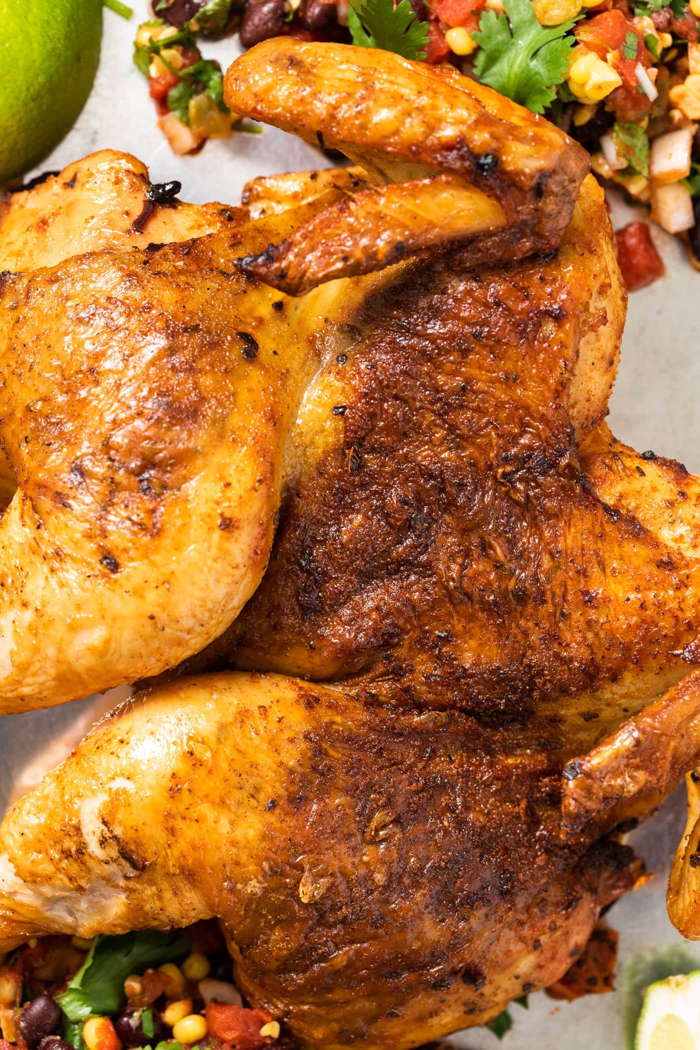 Close up top down view of spatchcocked chicken lying flat on steel pan after being grilled with salsa and limes around