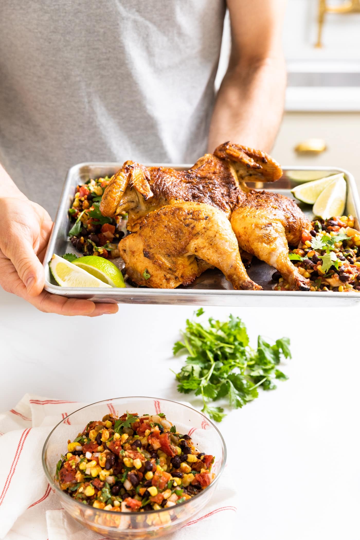 Two hands holding steel pan with an entire grilled chicken on it with limes and salsa around with extra cilantro