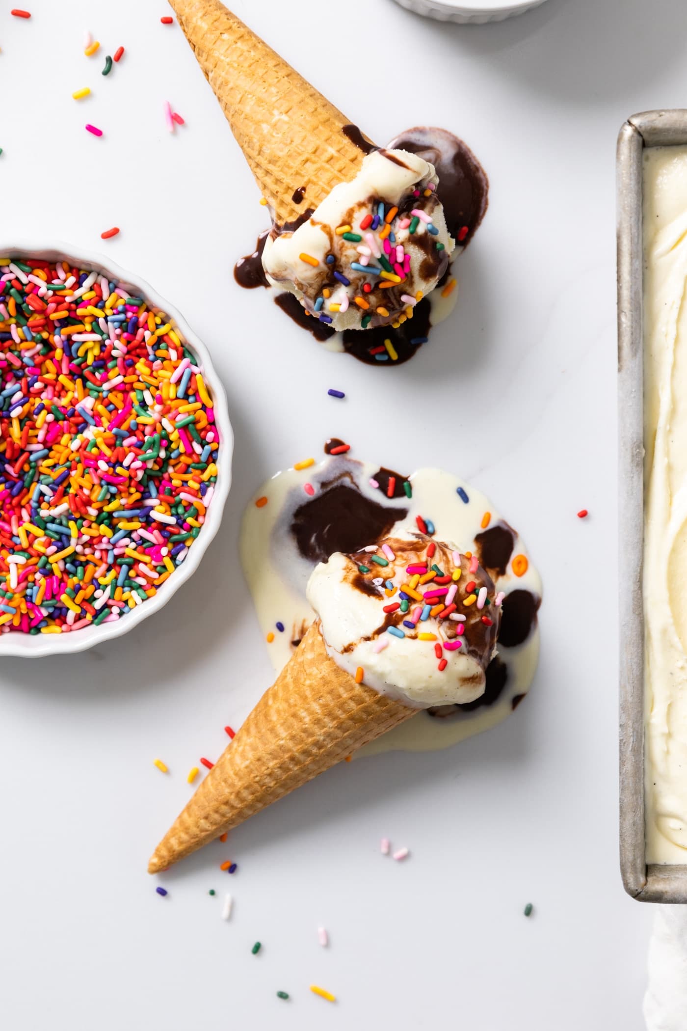 Top down view of vanilla ice cream sitting in ice cream cones with fudge and sprinkles all over the top
