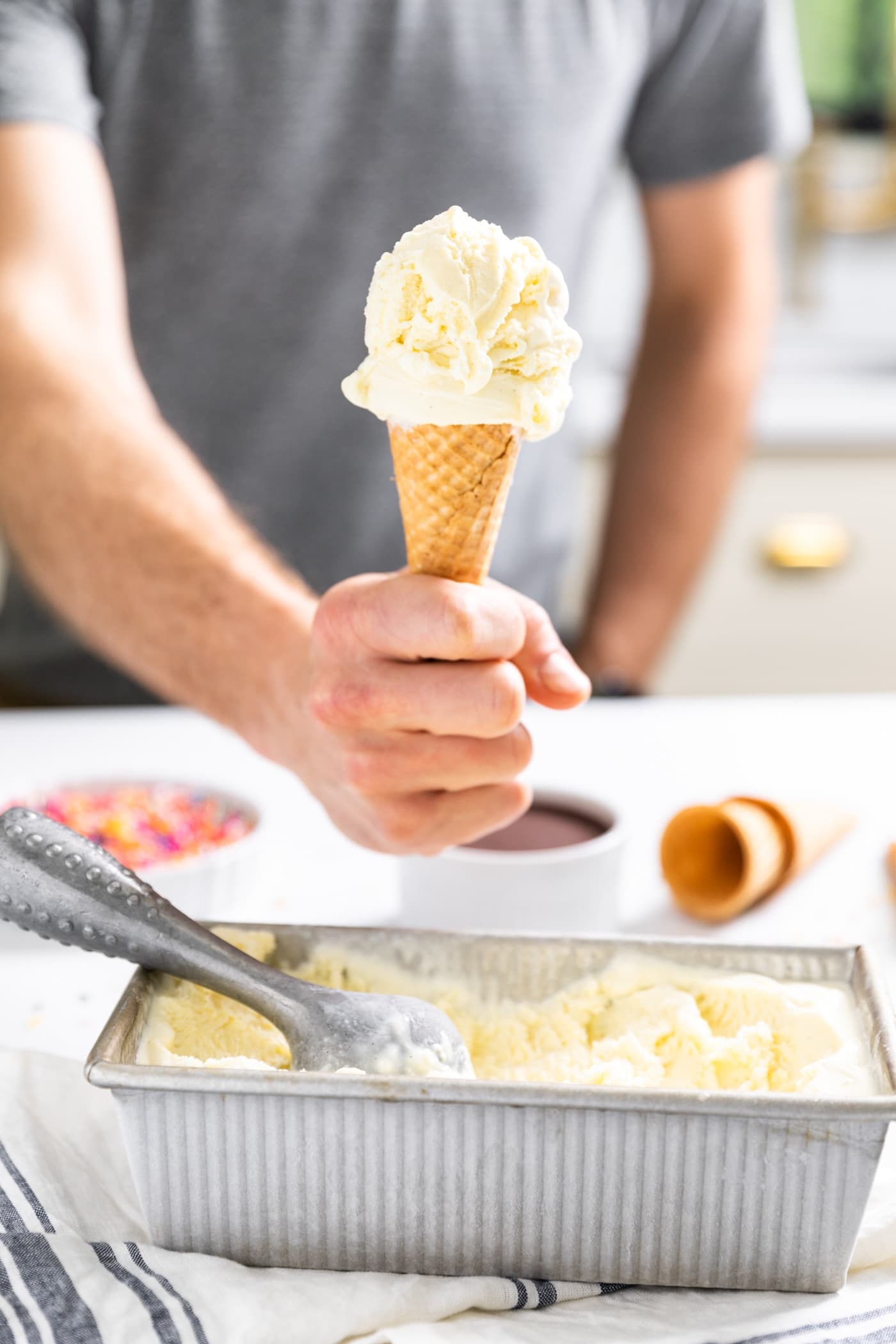 Hand holding ice cream cone filled with vanilla ice cream with rest of vanilla ice cream in silver container below sitting on countertop