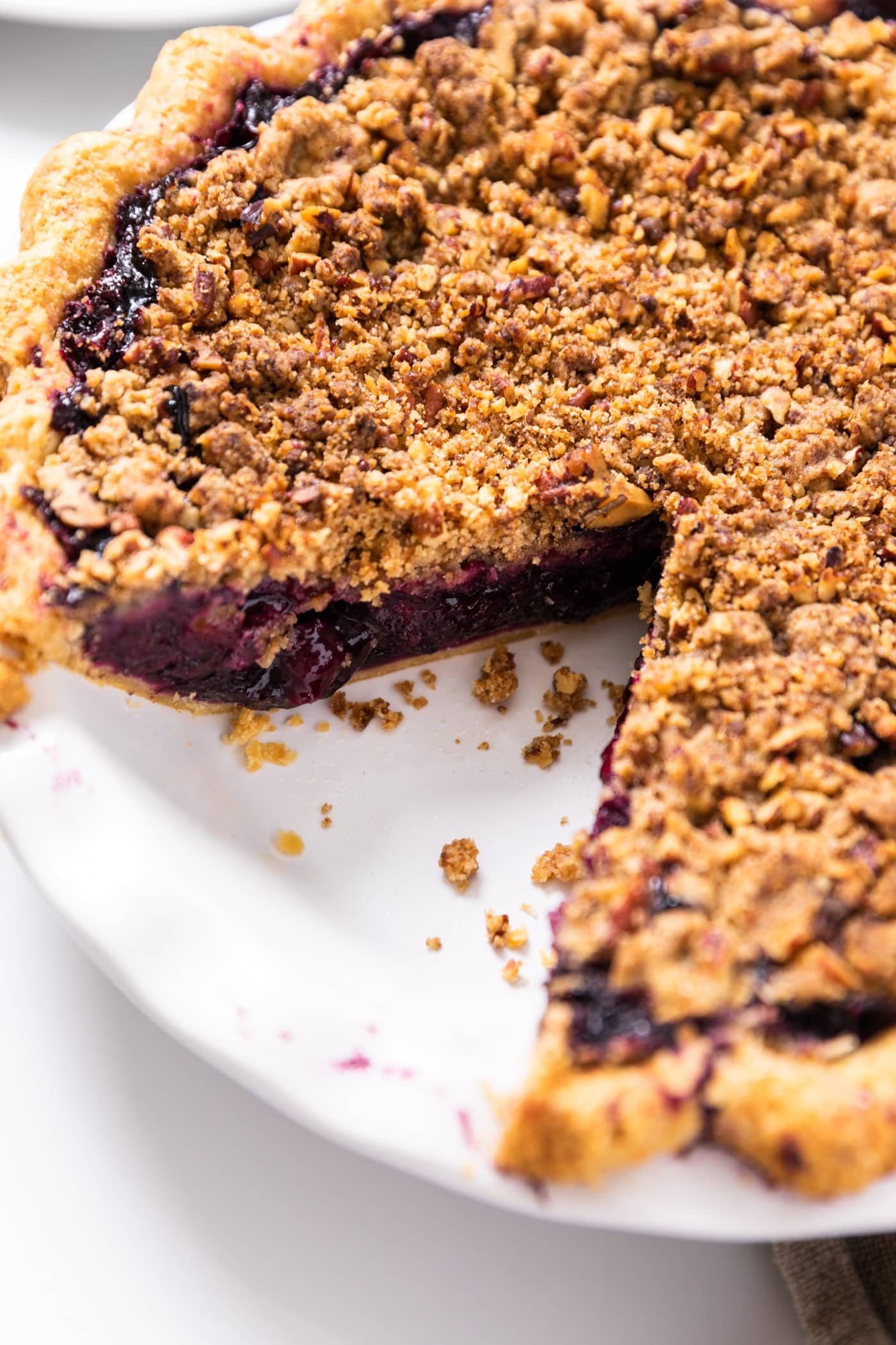 Blueberry pie sitting inn white baking dish with slice taken out showing the purplish inside of the pie with a streusel topping on top