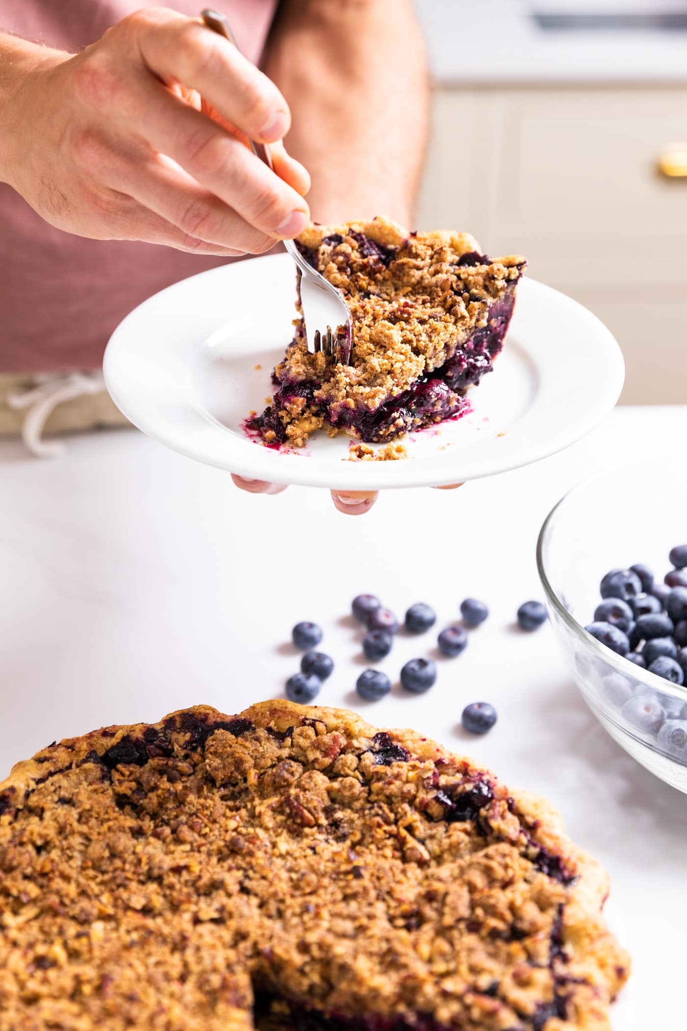One hand holding a plate with slice of pie with other hand holding fork taking piece from slice with rest of pie in the foreground