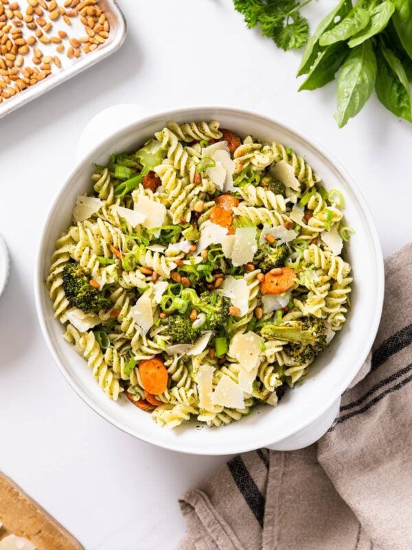 Top down view of white bowl filled with cooked pasta along with pieces of carrot and broccoli with extra herbs and pine nuts around