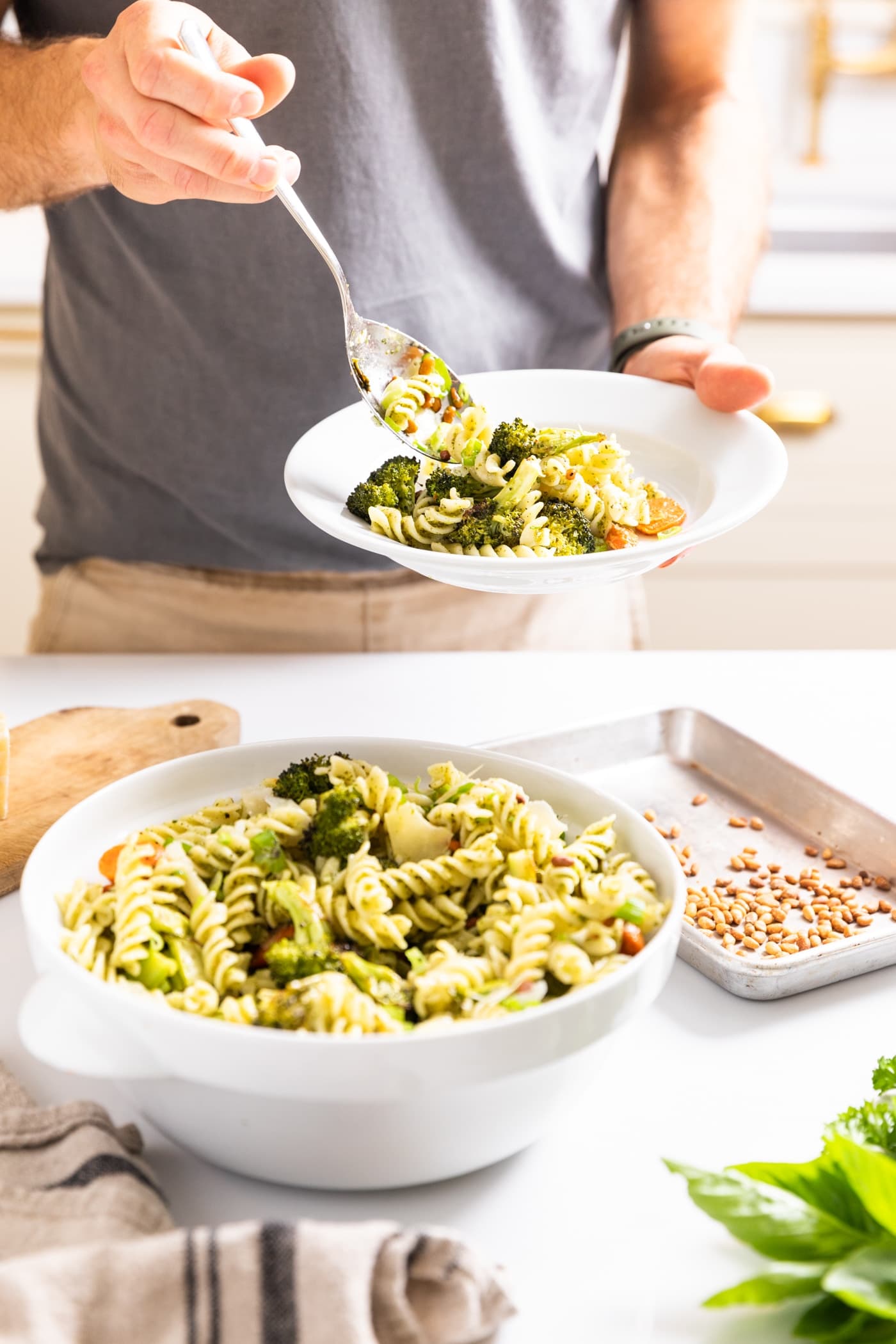 Hand holding white plate and scooping pasta salad onto plate from white bowl all on white countertop with tan cabinets in background