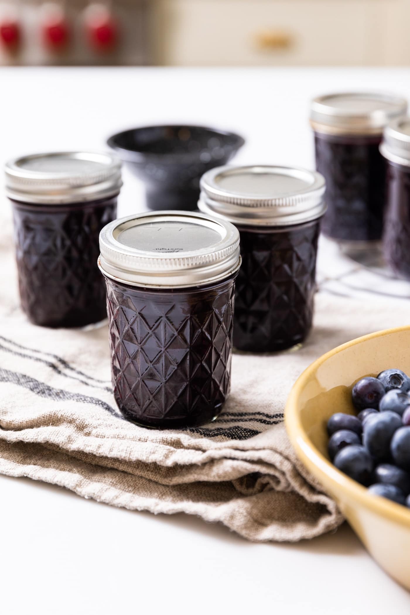 Glass jars with lids and rings on holding dark colored blueberry jam inside sitting on white countertop with bowl of blueberries in front
