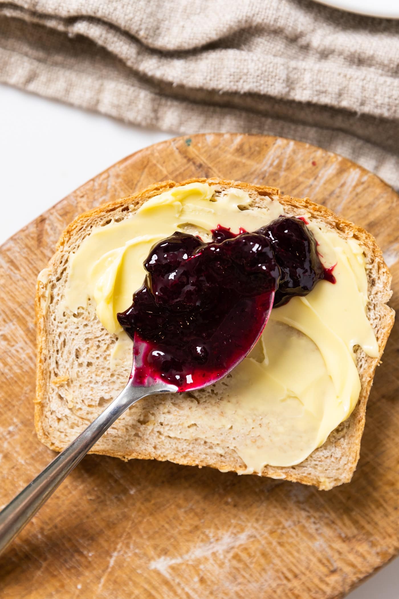 Spoon with purple colored blueberry jam sitting on buttered slice of brown bread on wooden cutting board with towel in background