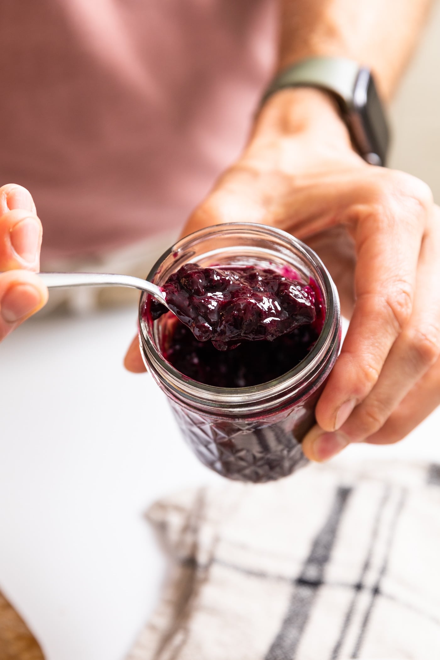 Hand holding glass jar filled with dark purple blueberry jam with other hand holding spoon scooping out some of the jam
