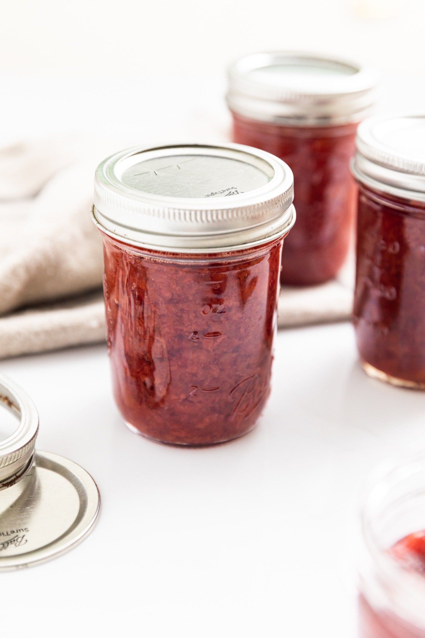 Three glass jars filled with red colored cherry jam sitting on white countertop with towel in background