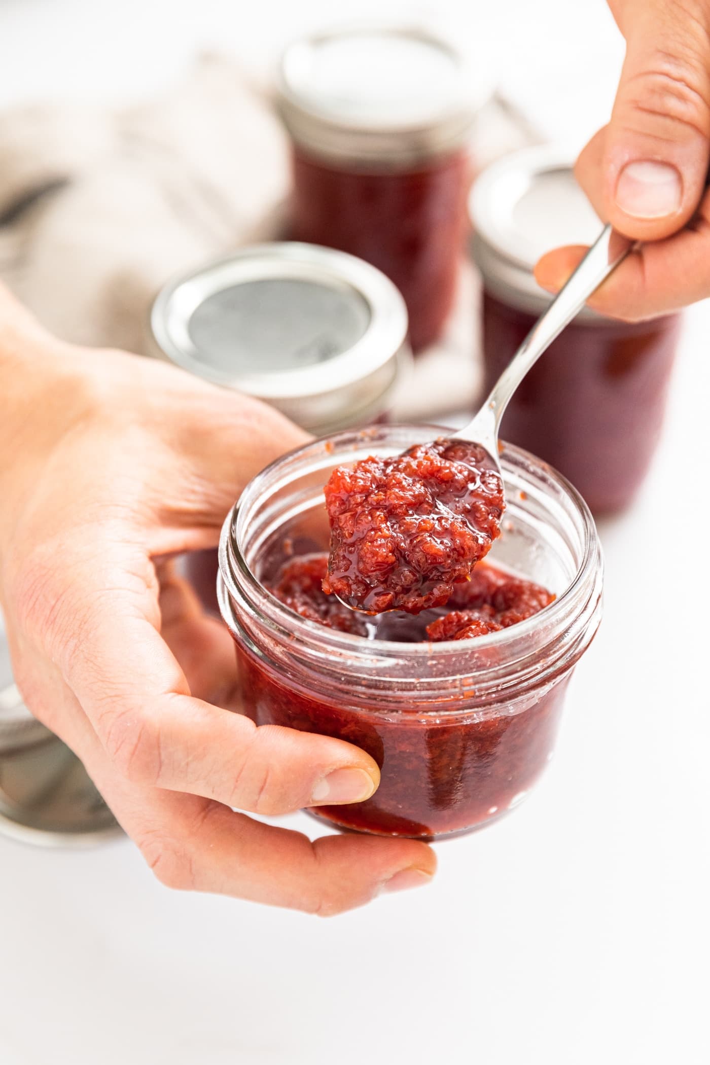 Close up view of two hands holding glass jar filled with red cherry jam with spoon holding some out to see the texture