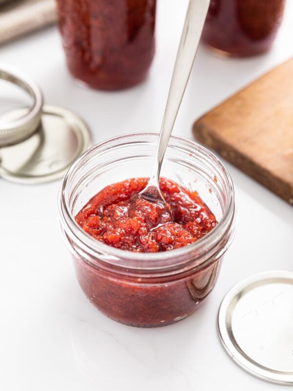 Small glass jar filled with red cherry jam with spoon sitting in it with other filled jars in background all on white countertop