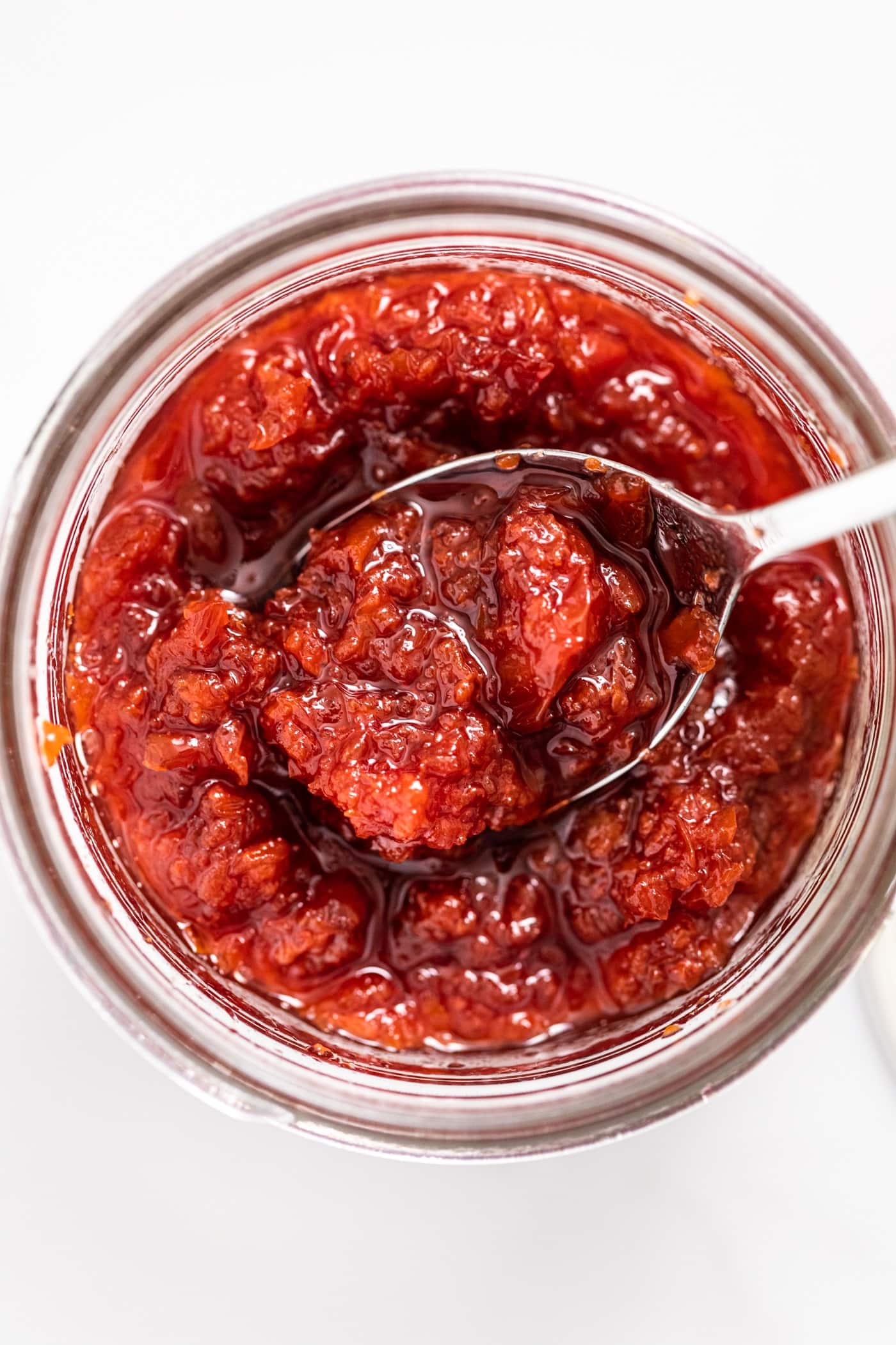 Top down view of red cherry jam with lots of texture sitting in glass container all on white countertop