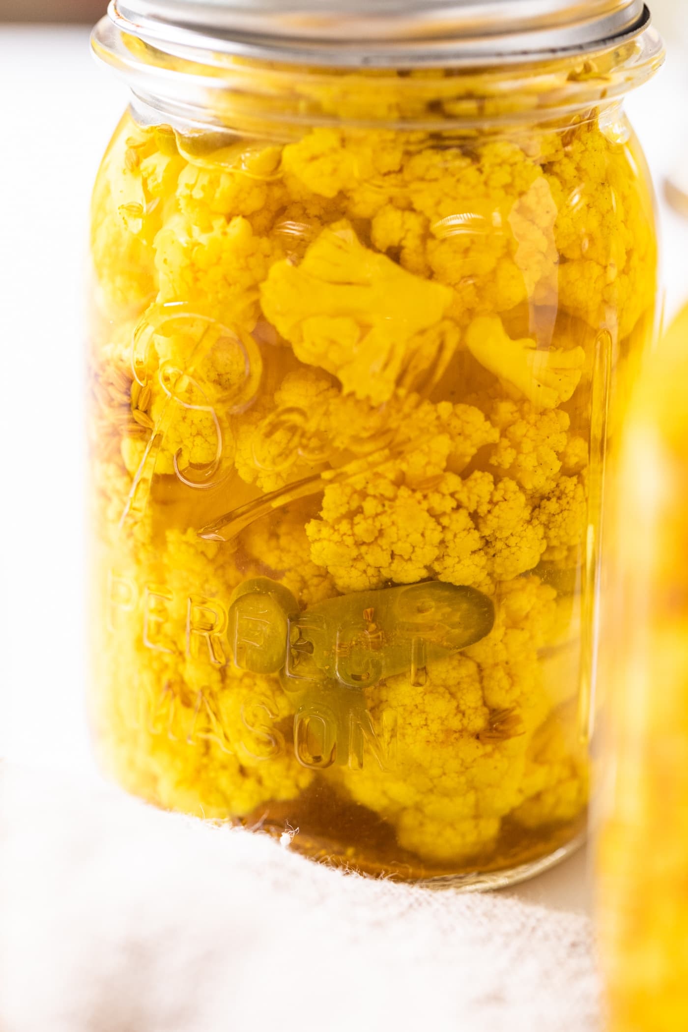Glass jar filled with yellow pickled cauliflower with slices of pepper showing all on white countertop