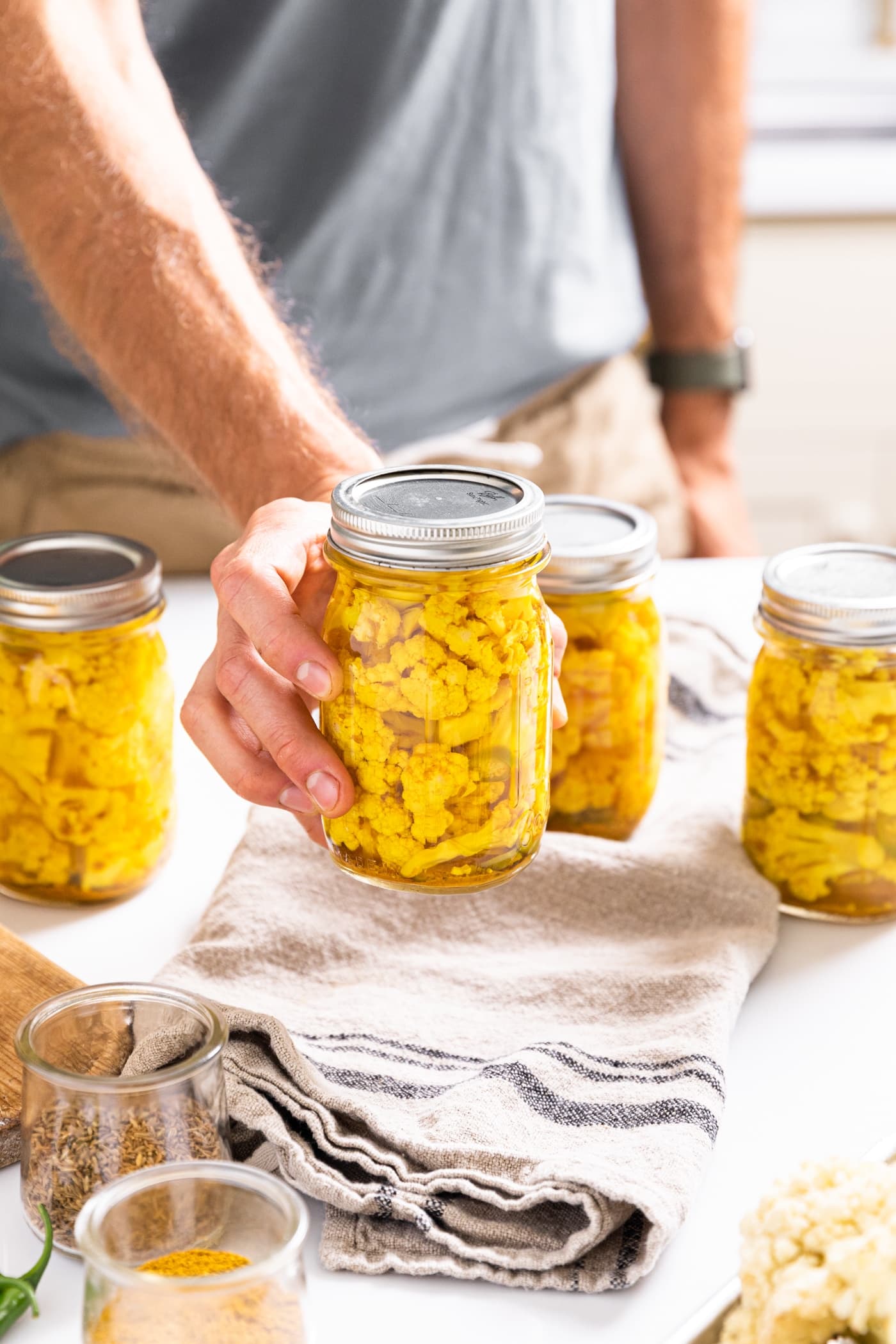 Hand holding glass jar filled with yellow pickled cauliflower with other filled jars in background all one white countertop with spices around