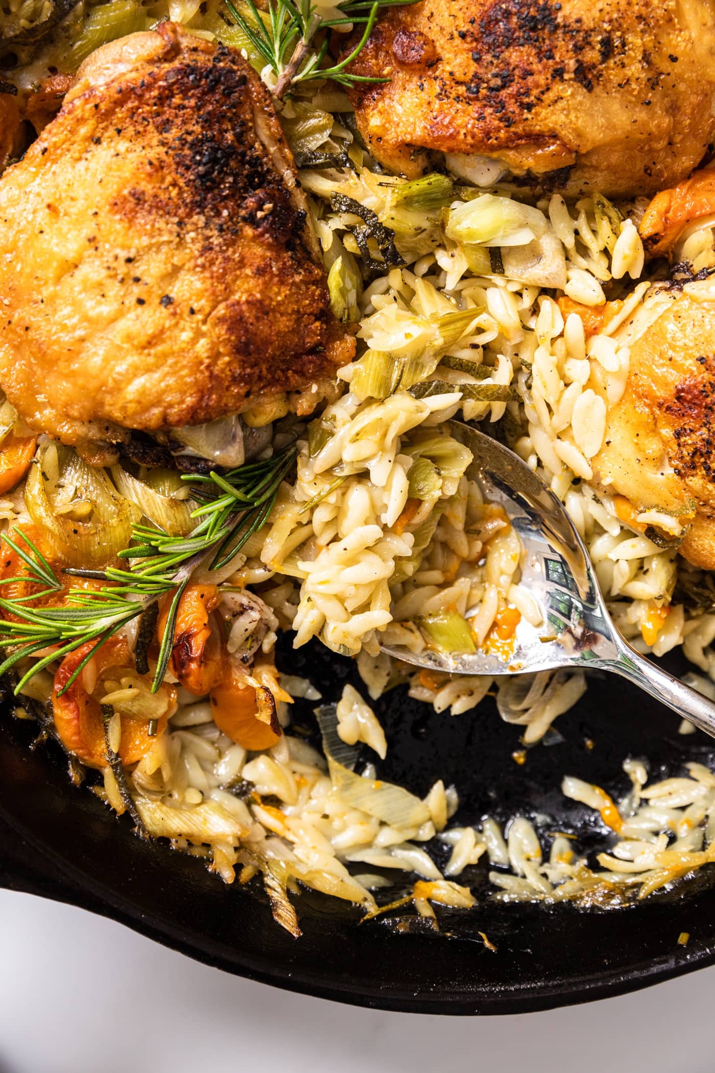 Top down view of spoon scooping out orzo from black skillet also filled with cooked chicken thighs