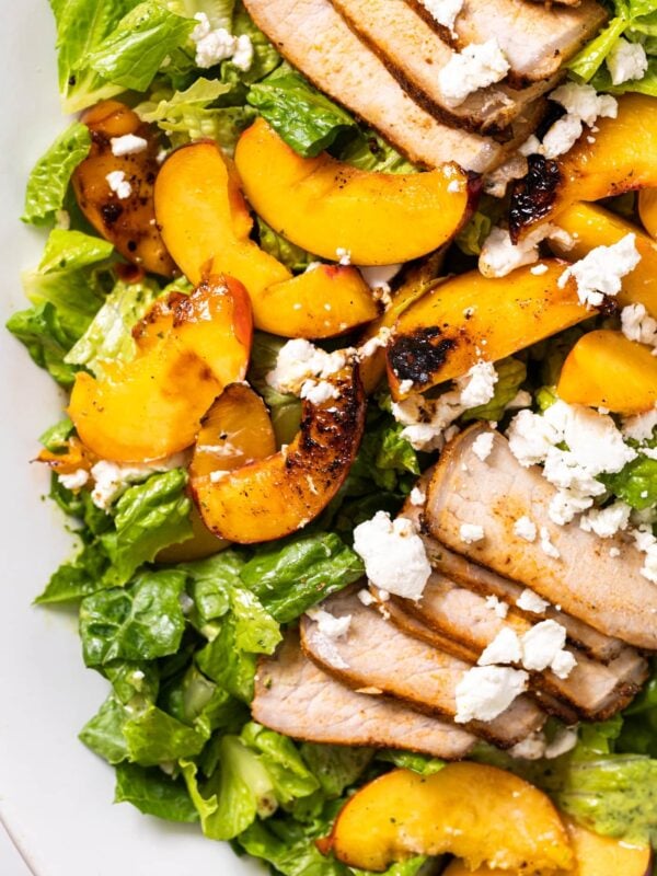 Top down view of white platter filled with slices of pork and nectarines topped with green dressing and pieces of white goat cheese