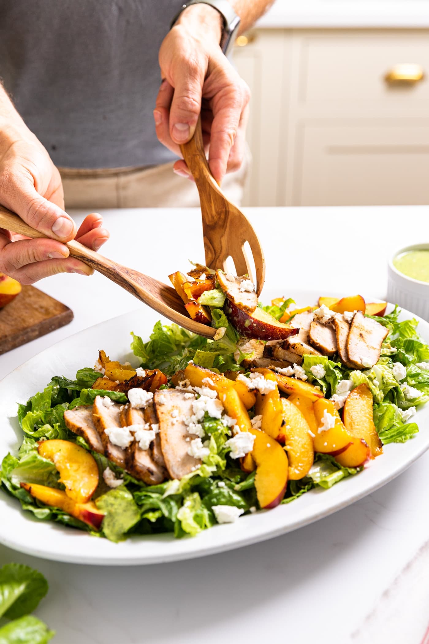 Two hands holding wooden tongs scooping up serving of salad from white platter filled with slices of nectarine and pork