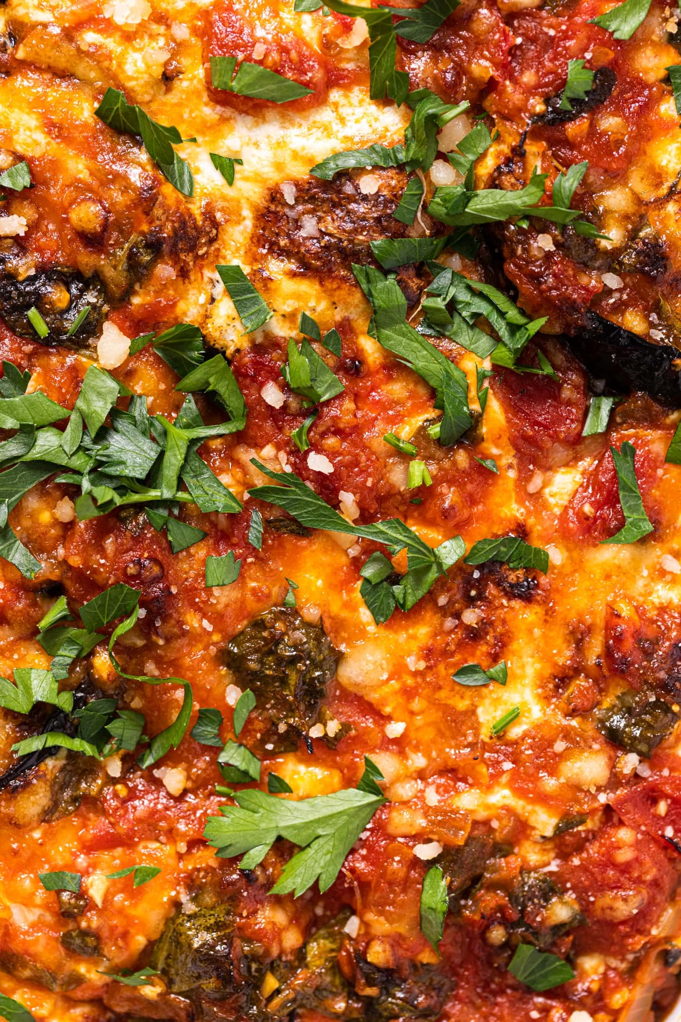 Close up view of the top layer of an eggplant lasagna recipe with bubbles of browned cheese and a fresh sprinkling of green herbs with bits of eggplant showing through the cheese