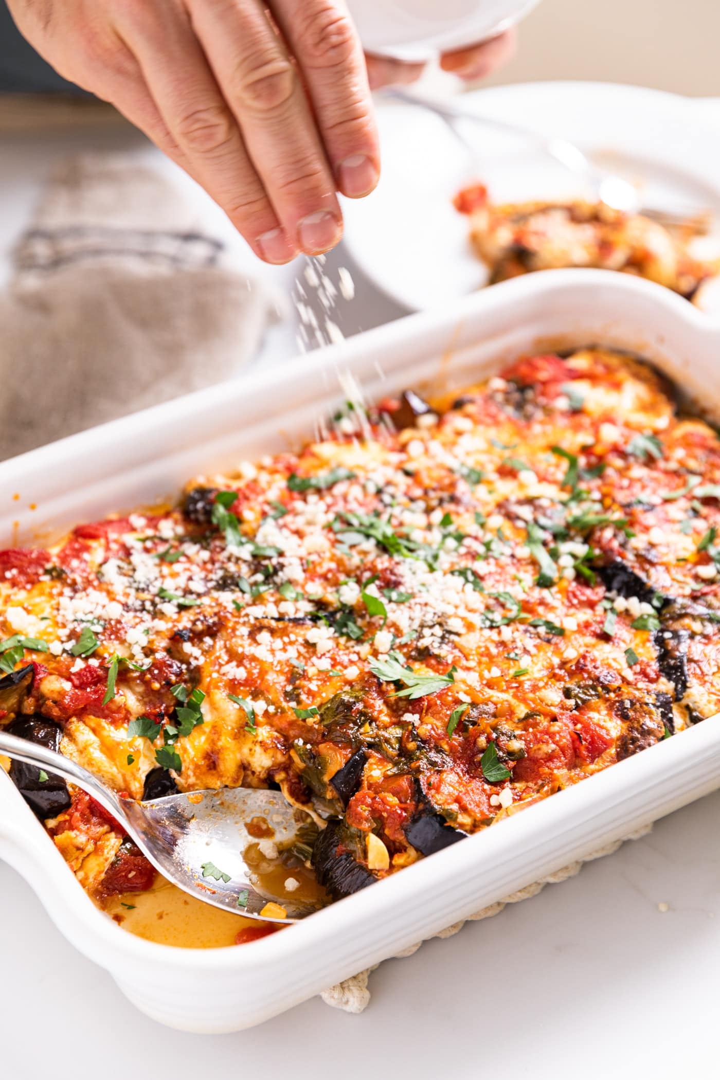 View of hand sprinkling Parmesan cheese bits on top of baking dish filled with eggplant parmesan with spoon at one end after slicing out a piece