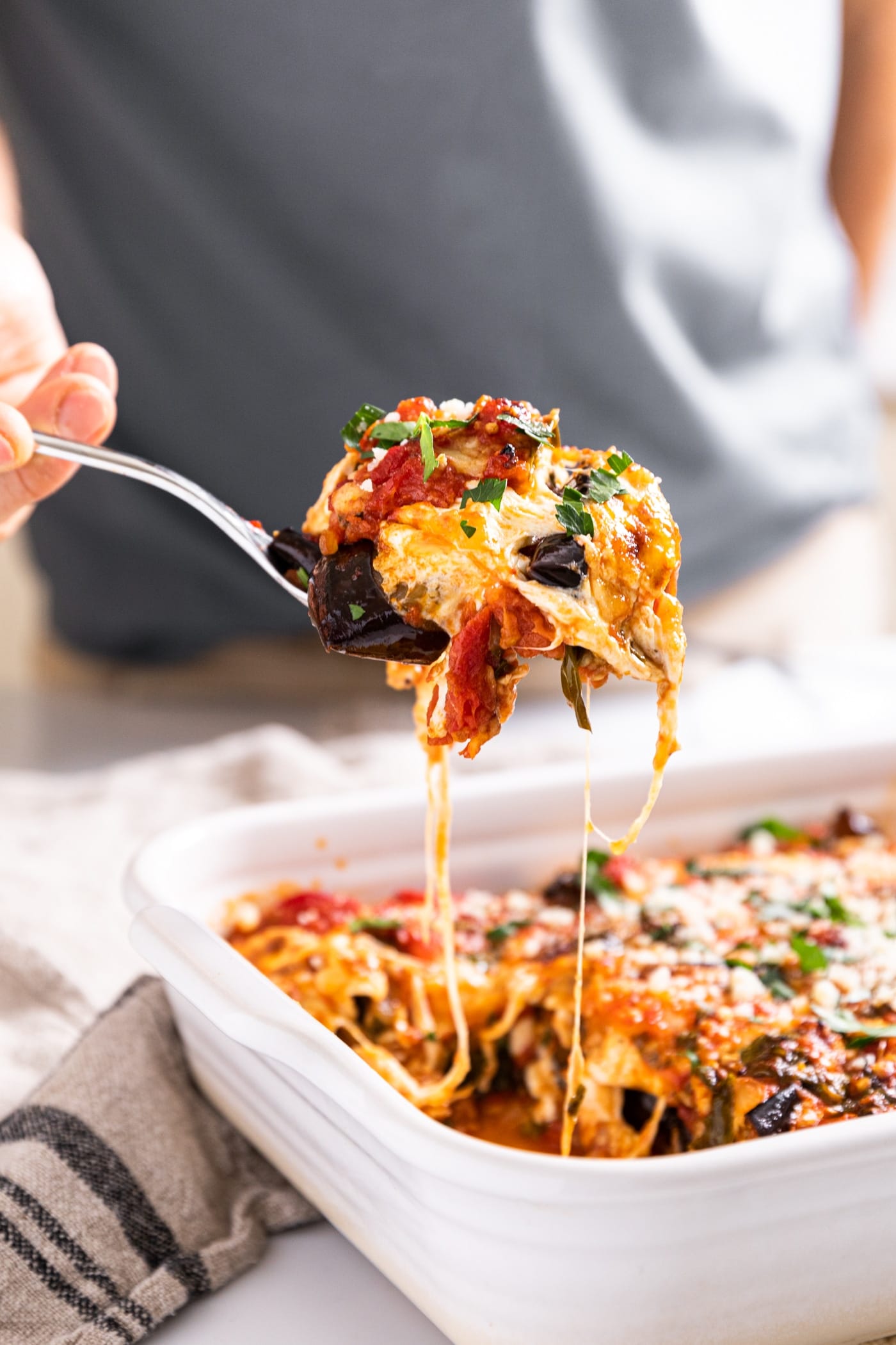 Hand holding scoop of eggplant parmesan from white baking dish after freshly being taken from the oven with strings of cheese still attached to rest of dish below