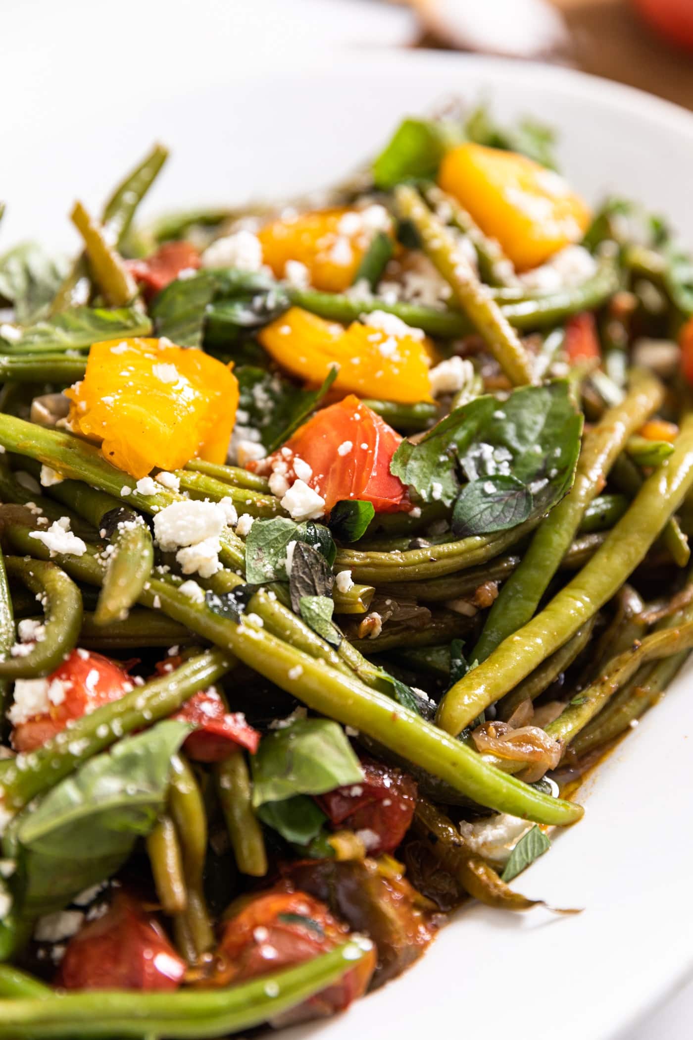 White platter filled with green beans and tomatoes and fresh herbs with feta cheese all over the top