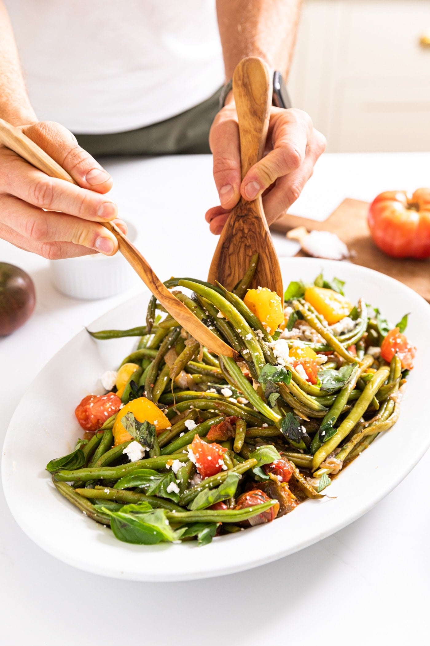 Two hands holding salad tongs pulling up serving of green beans and tomatoes from white platter sitting on white countertop with bright red tomato in background