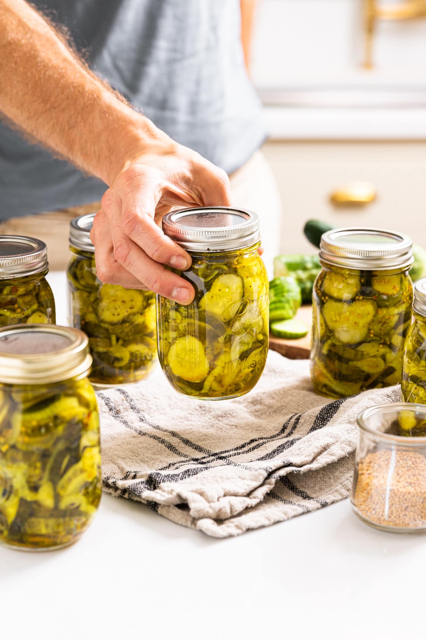 Hand holding glass jar filled with bread and butter pickles in the action of setting jar down on cloth with other glass jars all around