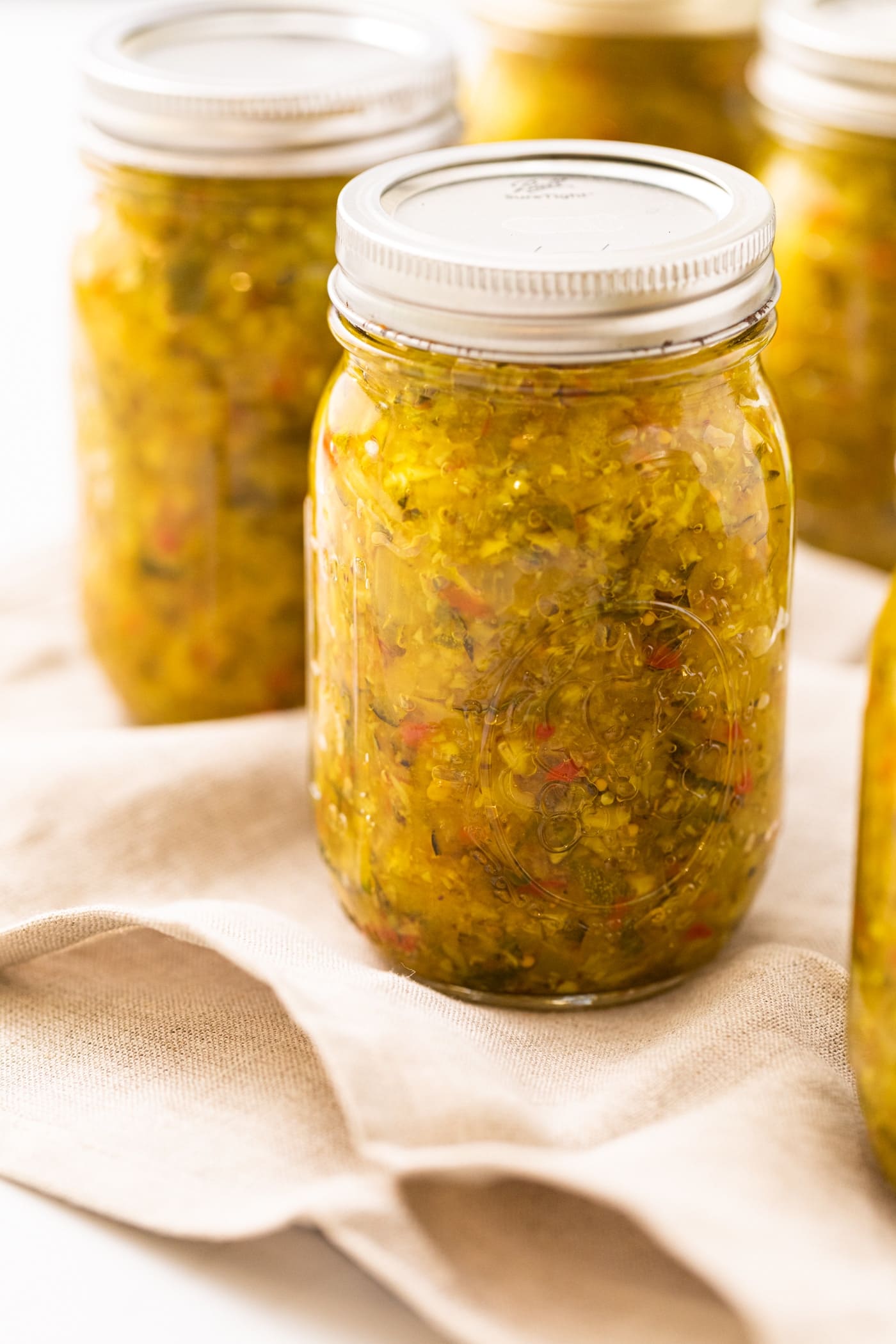 Glass jar filled with yellow colored pickle relish sitting on taupe cloth on white countertop